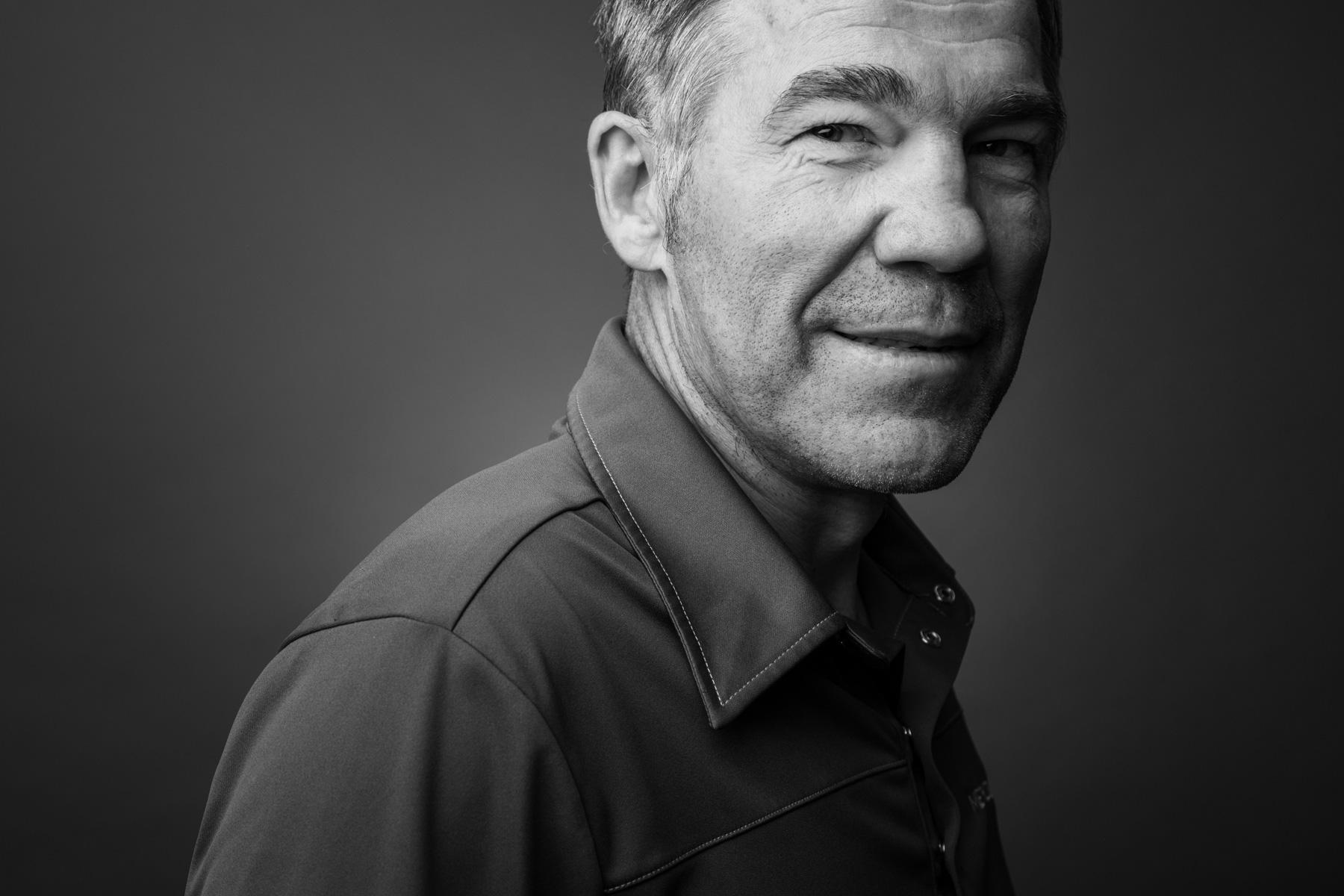 Black and white portrait of a middle-aged man smiling, wearing a collared shirt against a plain dark background.
