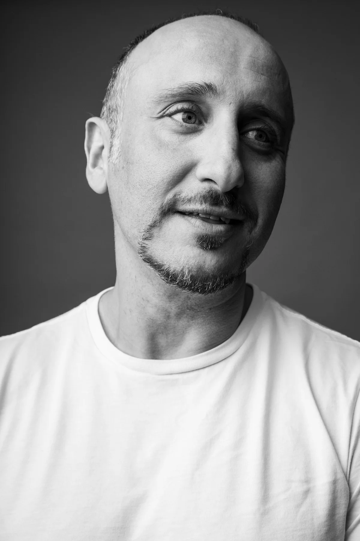 Black and white portrait of a man with a short beard and mustache, bald head, wearing a white t-shirt, looking to the side with a slight smile.
