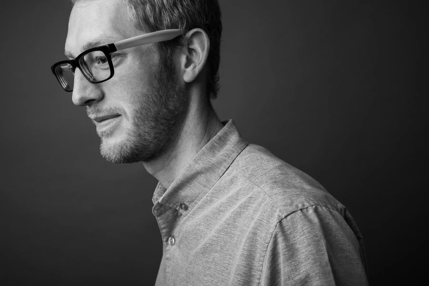 Black and white profile of a young man with glasses, beard, wearing a collared shirt, looking down against a dark background.