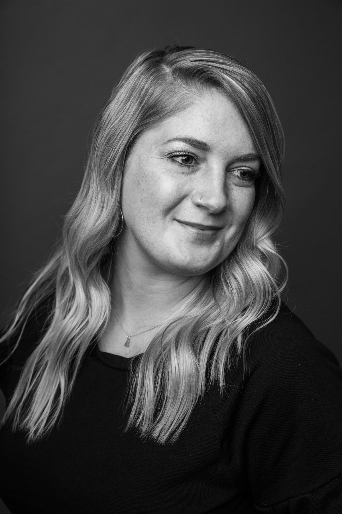 A young woman with wavy hair, wearing a black top and a necklace, looking down and smiling slightly in a black and white portrait against a dark background.