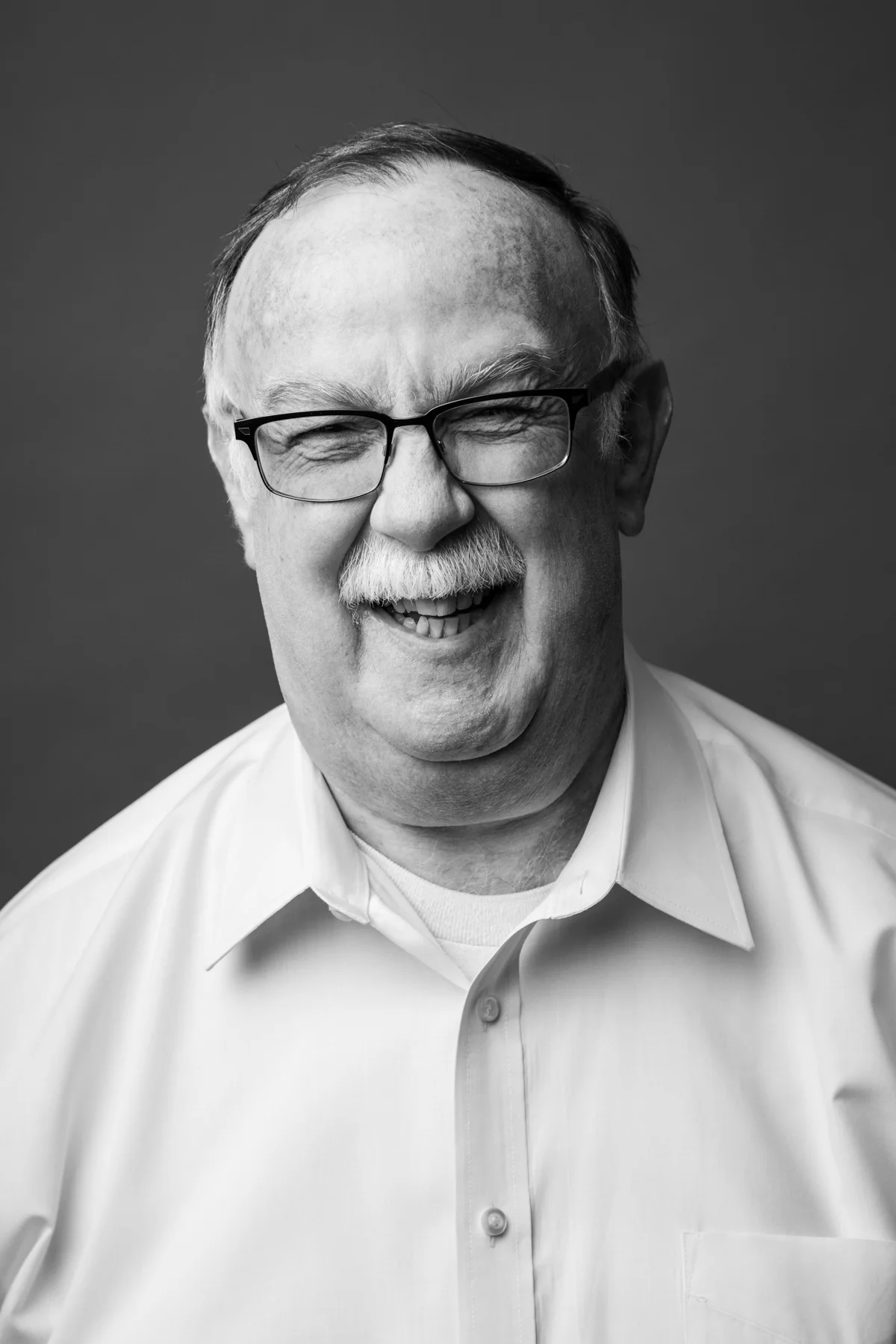 Black and white portrait of a smiling man with glasses, a mustache, wearing a dress shirt.