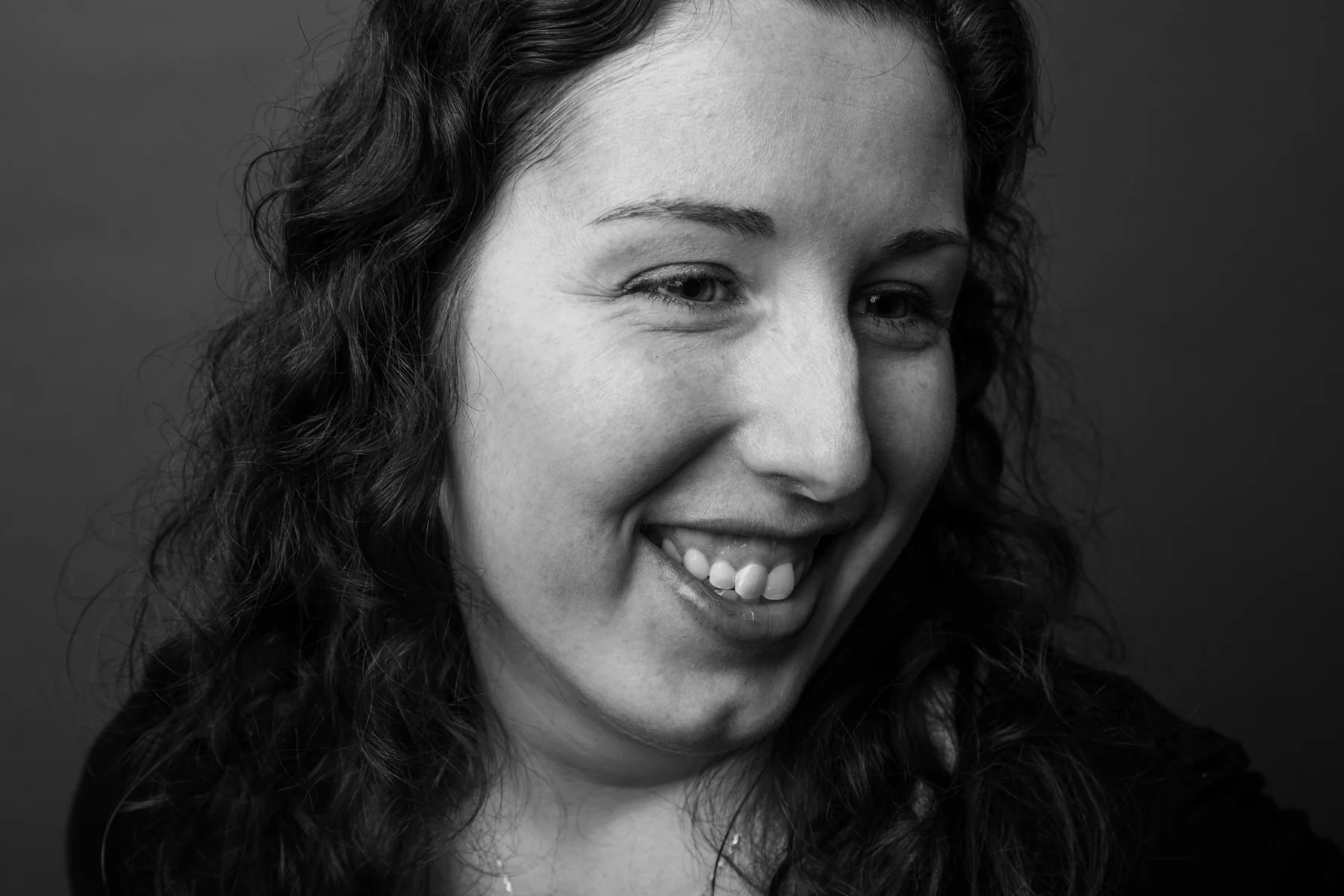 Black and white close-up portrait of a smiling woman with curly hair, showing her teeth and dimples.