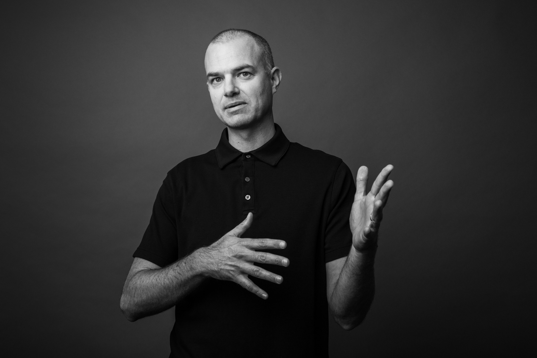 Black and white portrait of a man with a shaved head, wearing a dark polo shirt, gesturing with one hand on his chest and the other raised, against a plain background.