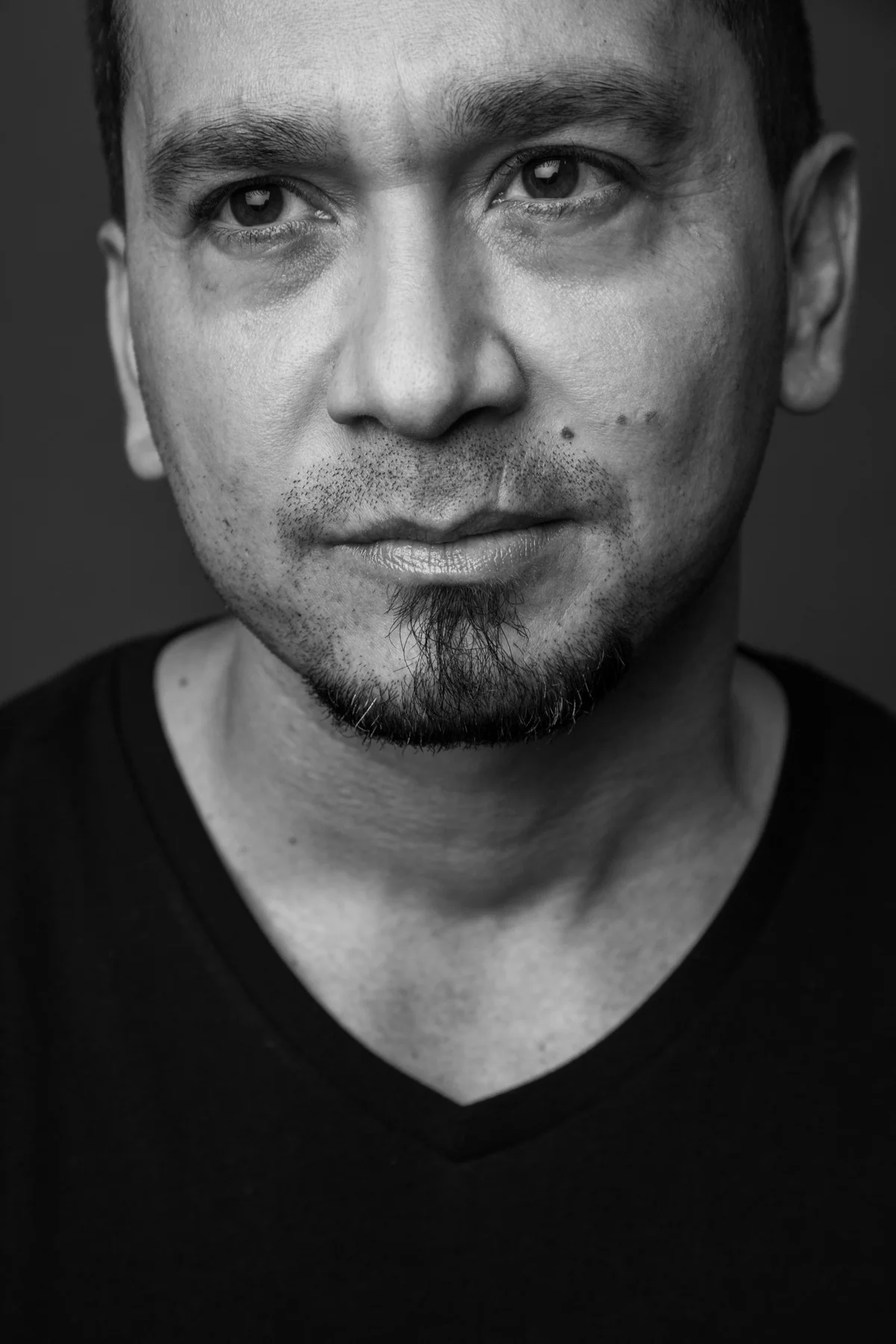 Black and white close-up portrait of a man with short hair, facial hair, and wearing a black V-neck shirt.
