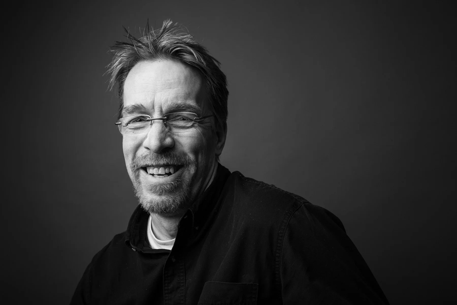Black and white portrait of a smiling man with glasses, spiky hair, and a goatee, wearing a dark shirt, against a dark background.