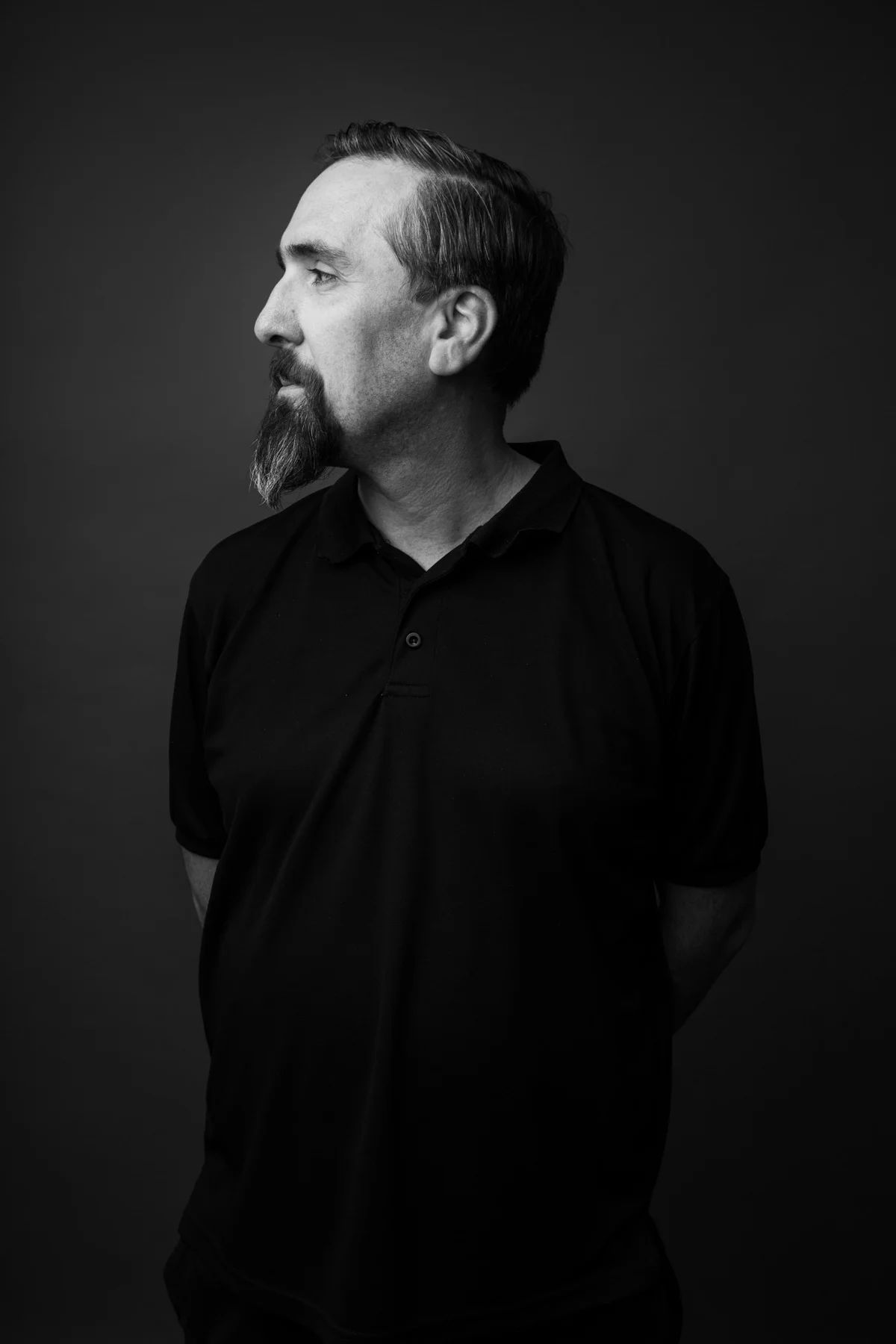 A black and white portrait of a man with a beard, looking to the left, wearing a black collared shirt, standing against a dark background.