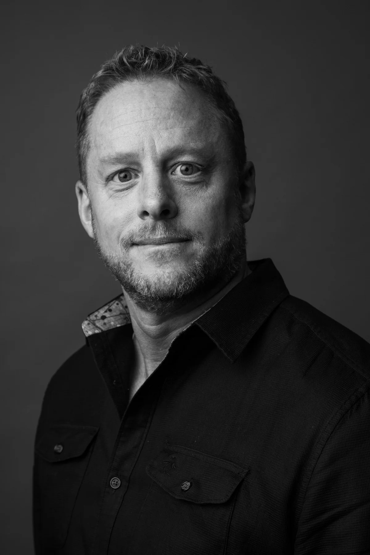 Black and white portrait of a smiling man with short hair and a beard, wearing a dark shirt with patterned collar details, in a studio setting.