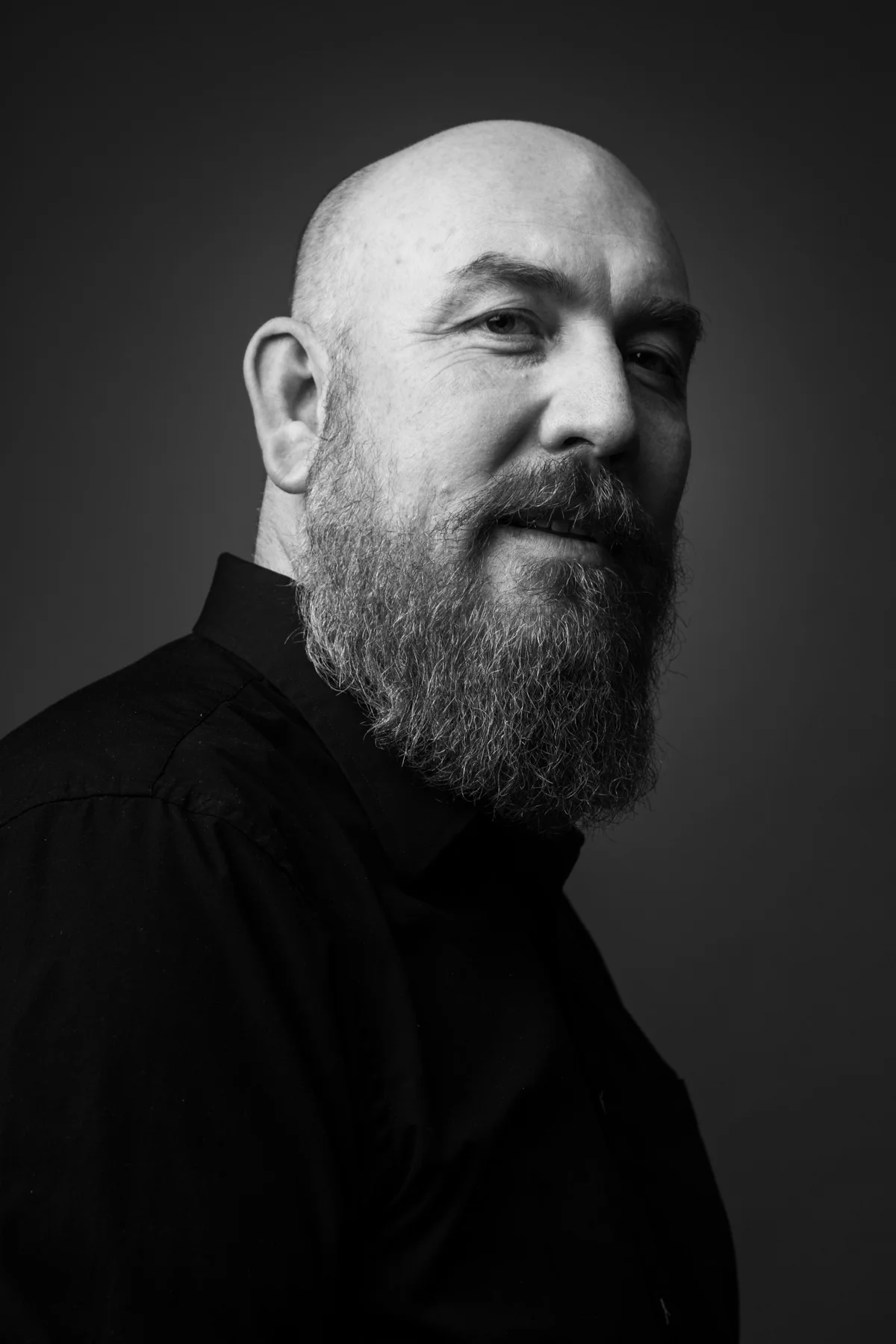 Black and white portrait of a bearded man with a shaved head, wearing a dark shirt, smiling slightly, against a dark background.