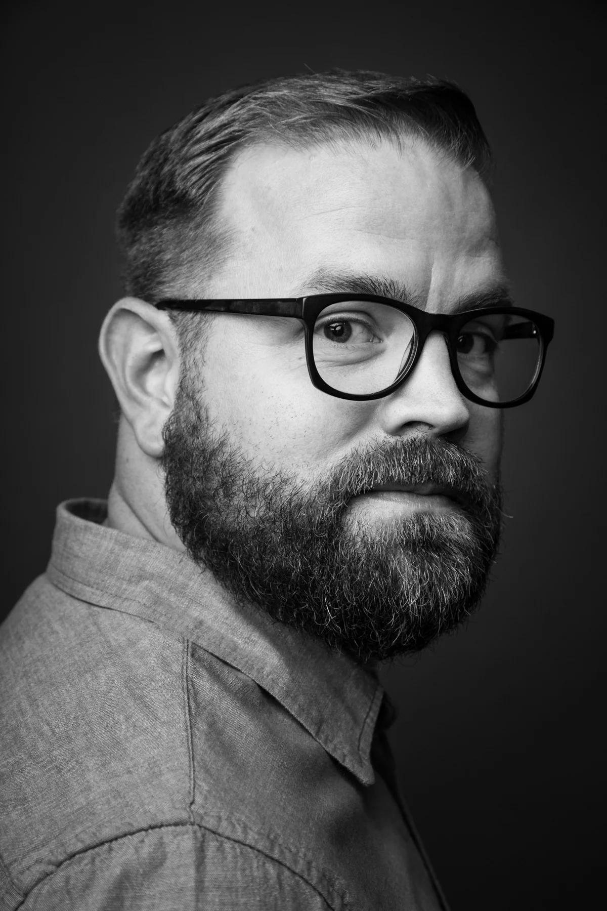 Black and white portrait of a man with glasses and a beard, looking over his shoulder at the camera.