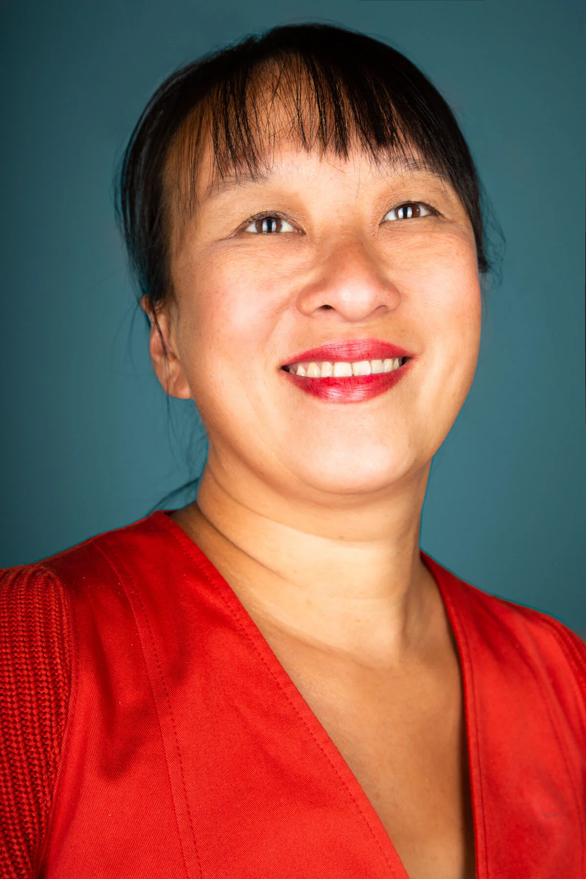 A smiling woman with dark hair wearing a red top against a teal background.