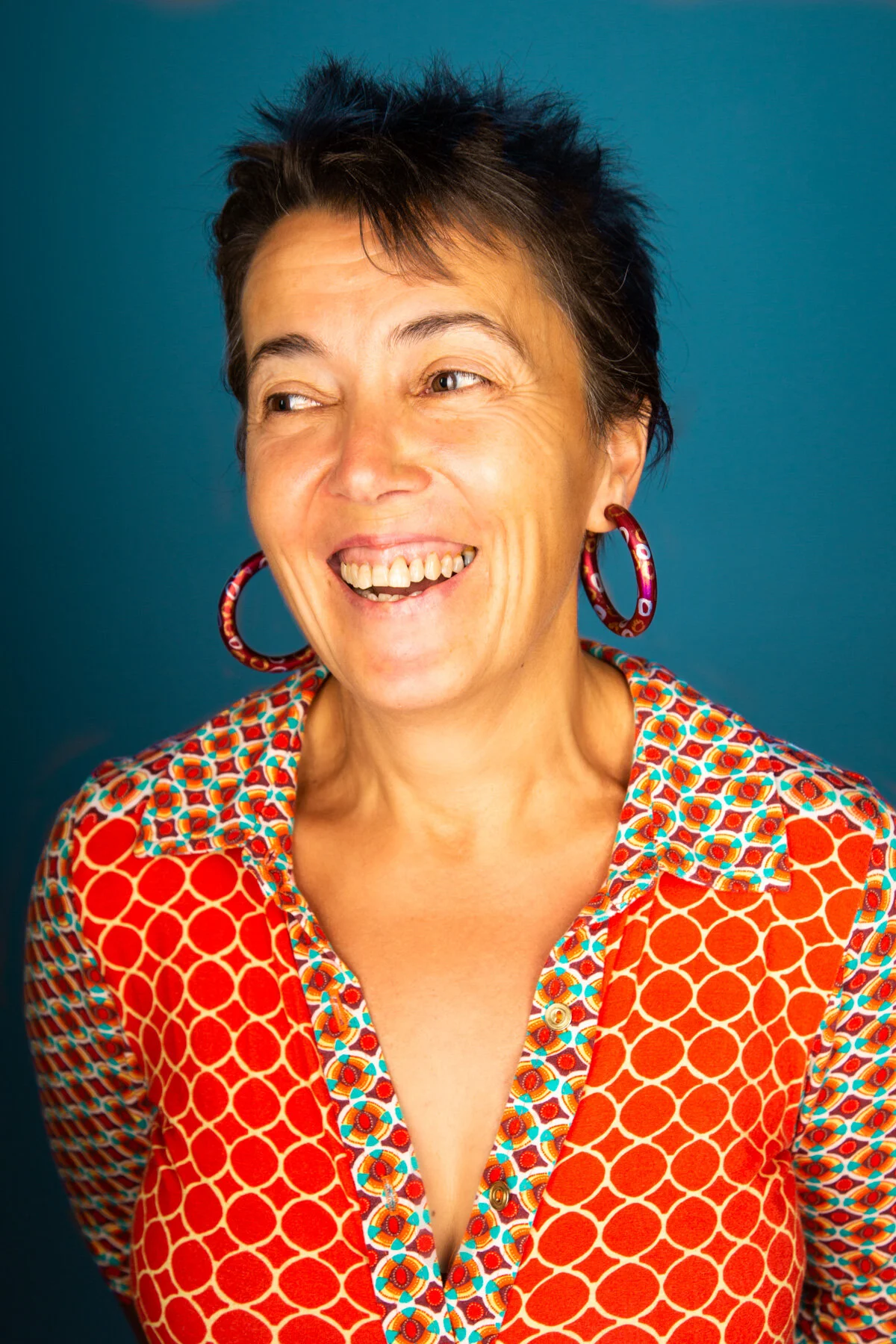 A woman with short dark hair, smiling and looking slightly to the side, wearing large colorful earrings and a vibrant red patterned shirt against a solid blue background.