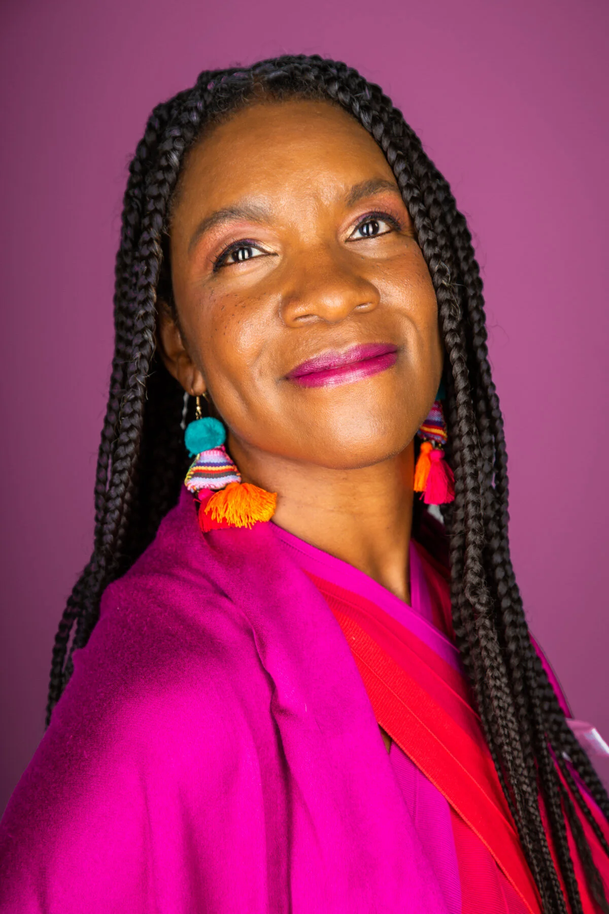 A woman with long braided hair, colorful earrings, and pink clothing, posing against a purple background.