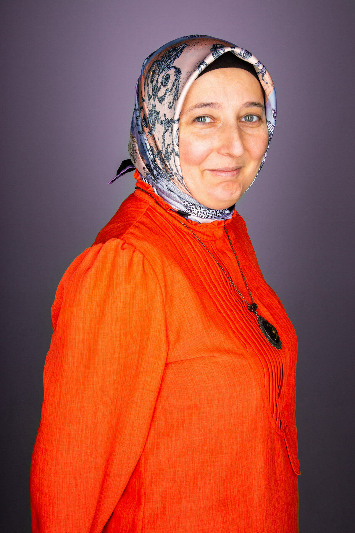 A woman with light skin wearing a patterned headscarf, an orange blouse, and a necklace with a dark pendant, standing against a neutral background.