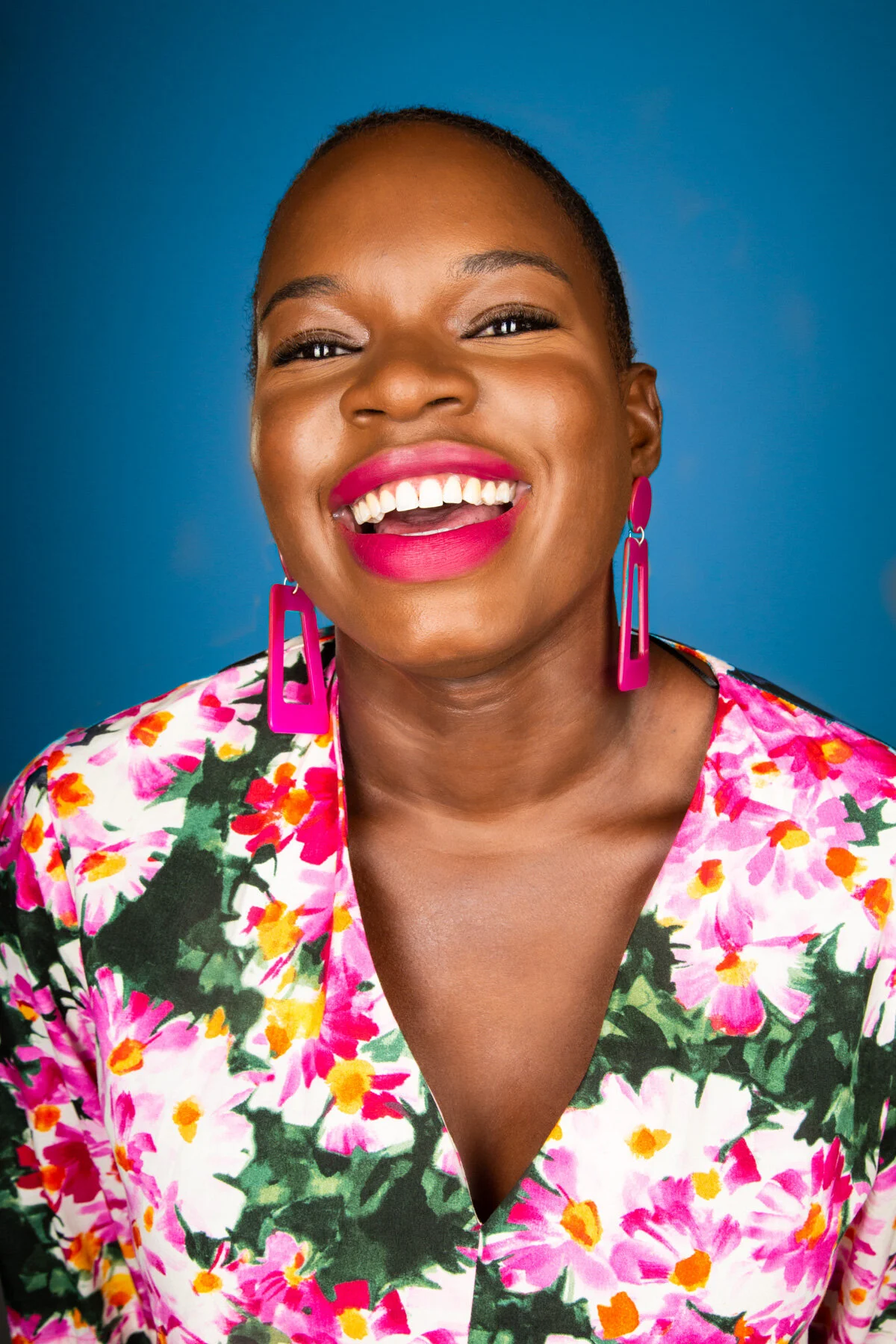 A woman smiling broadly, wearing bright pink lipstick, large pink earrings, and a colorful floral top against a blue background.