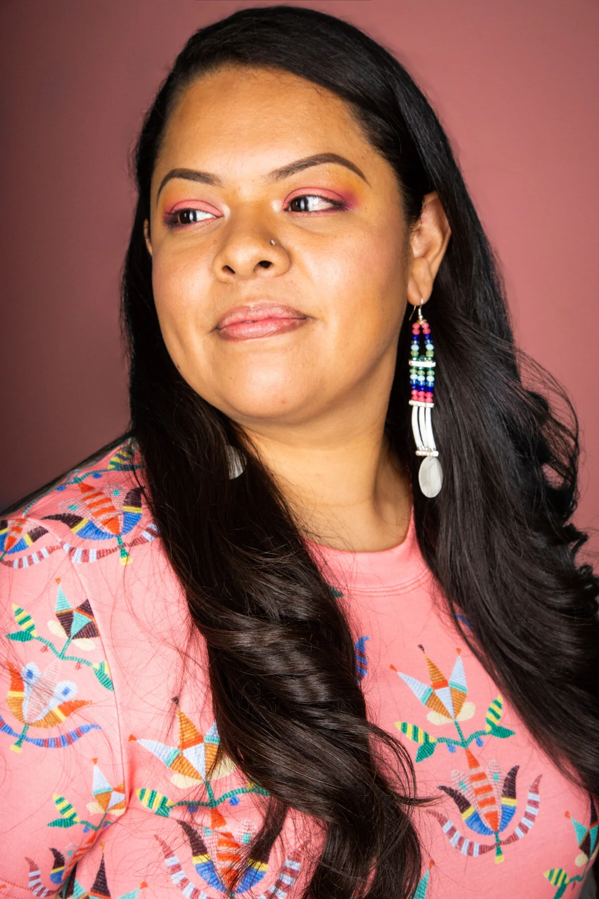 A woman with long dark hair wearing a pink shirt with colorful embroidery and colorful beaded earrings. She has a nose piercing and pink eye makeup, looking slightly to her right against a pink background.