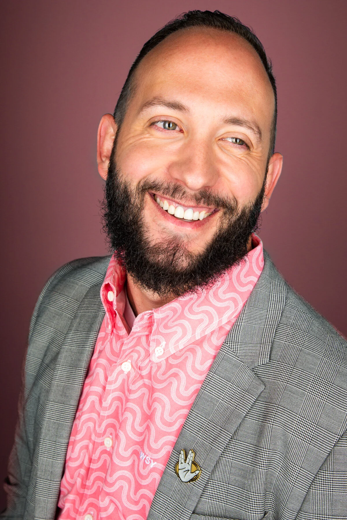 A smiling man with a beard and short hair wearing a pink patterned shirt, a gray plaid blazer, and a lapel pin with a hand making a peace sign, posed against a pink background.
