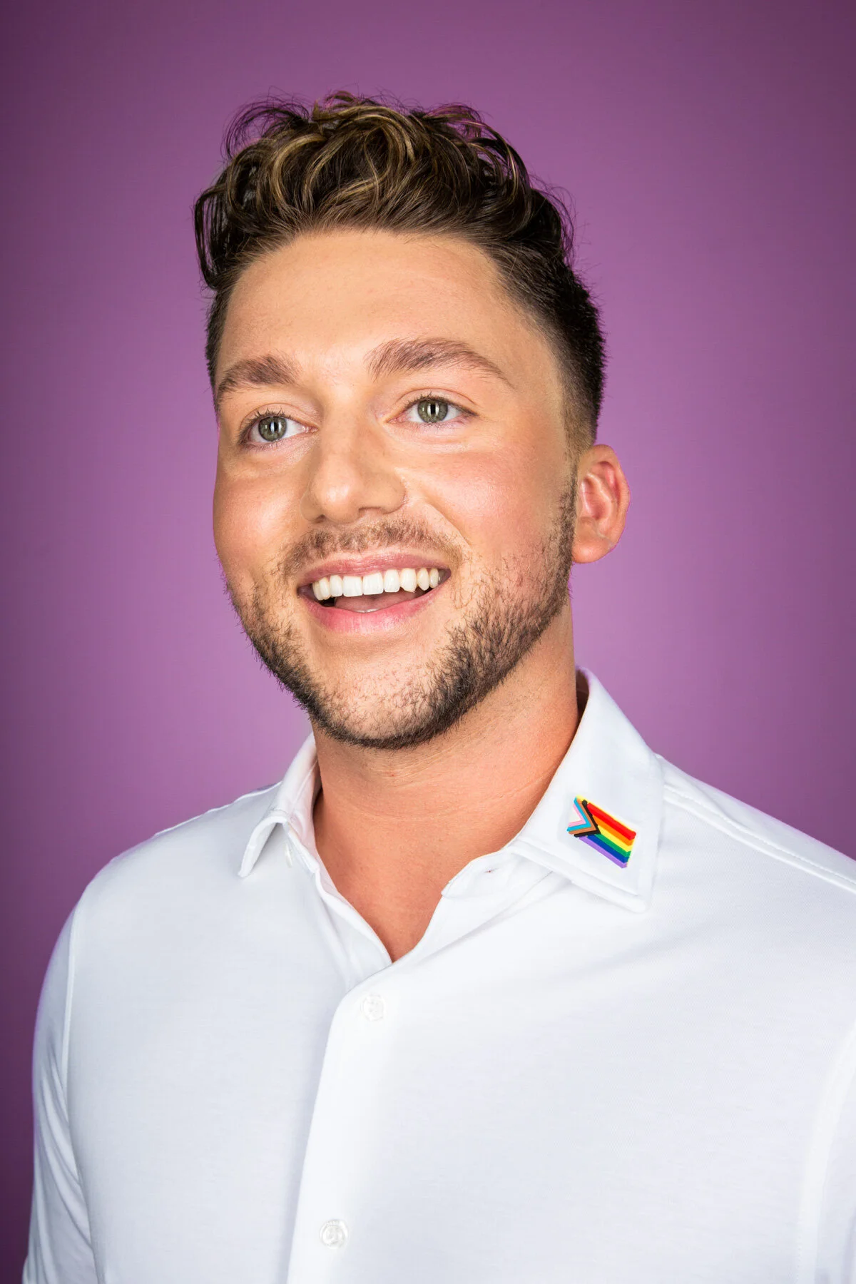A smiling man with short, dark brown hair, a beard, and blue eyes wears a white button-up shirt with a rainbow pride flag pin on the collar, against a purple background.