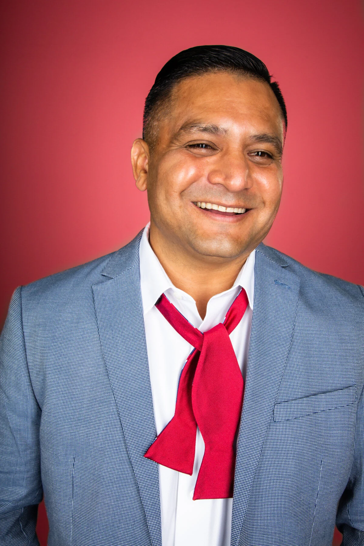 Smiling man in a blue suit with a white shirt and red tie, posing against a pink background.