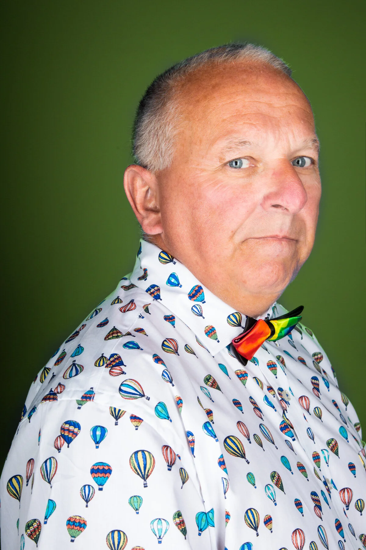 Close-up of a middle-aged man with light skin, short gray hair, blue eyes, wearing a white shirt with colorful hot air balloon patterns and a rainbow-colored bow tie, against a green background.