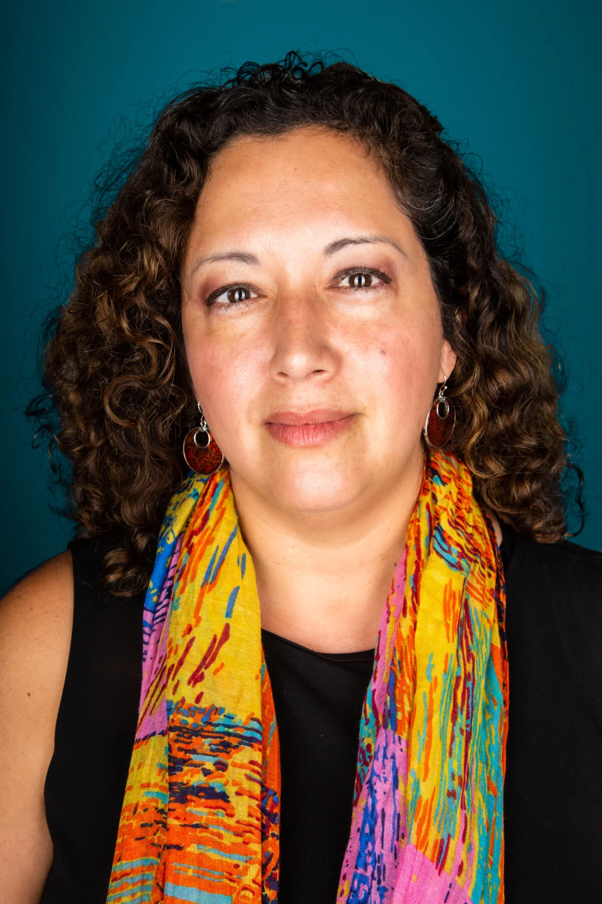 A woman with curly brown hair, wearing earrings, a colorful scarf, and a black top, posing against a teal background.
