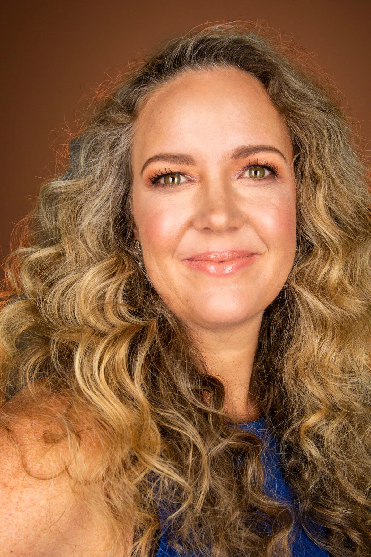 A woman with long, curly blonde hair looking at the camera with a soft smile, wearing a blue top and earrings, against a warm brown background.