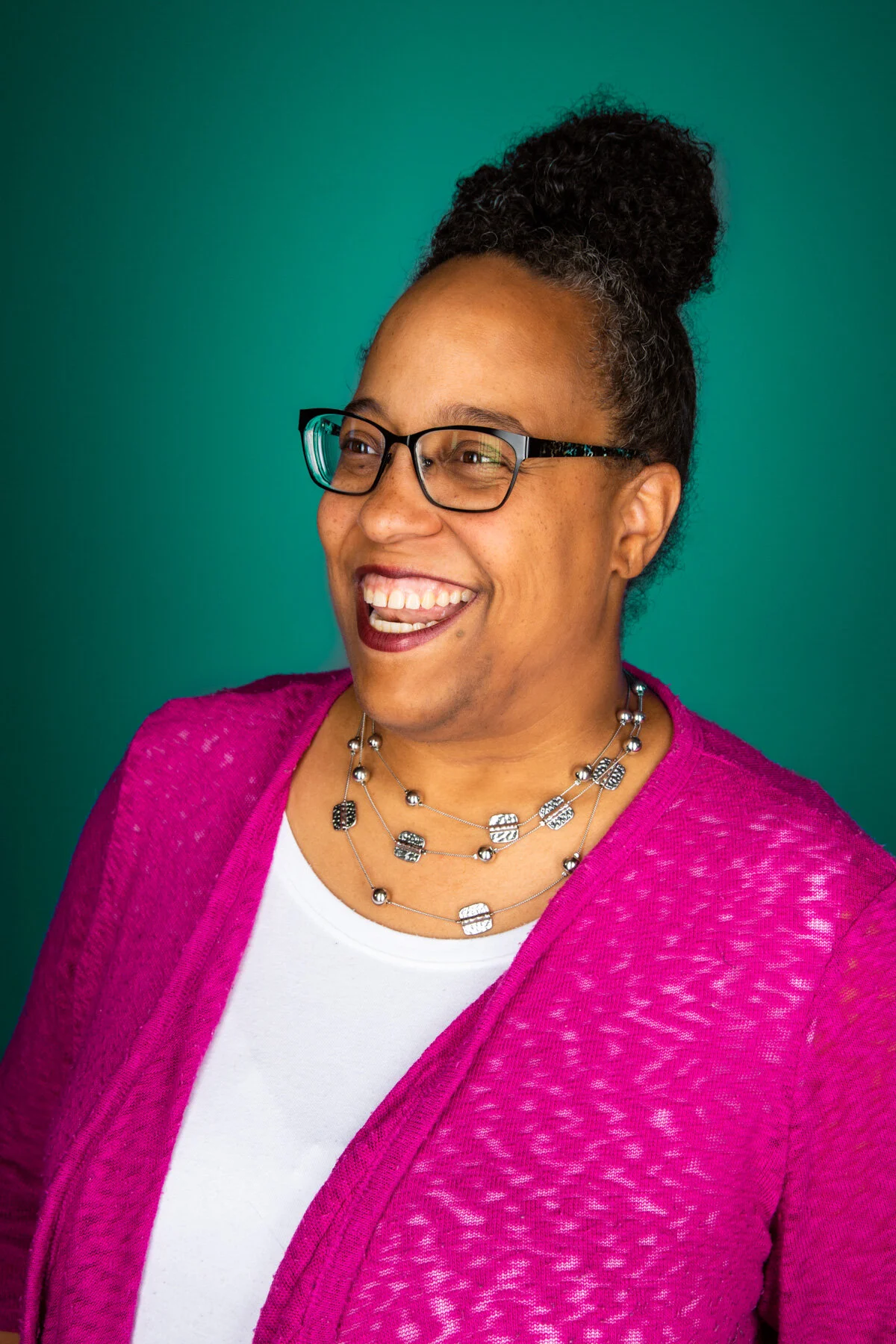 A woman with glasses wearing a pink cardigan and layered necklaces, smiling and looking to the side against a teal background.