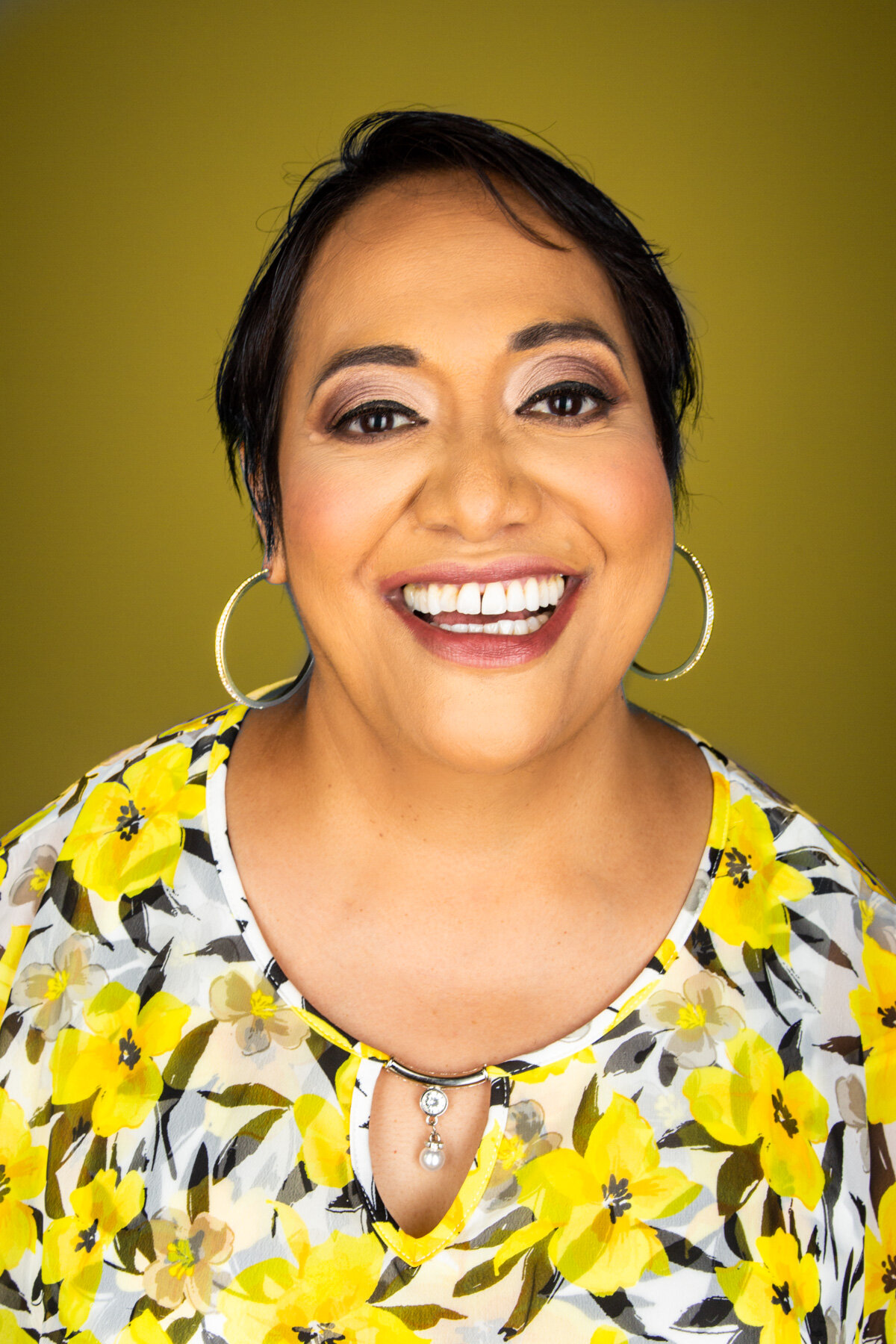 Smiling woman with short dark hair, hoop earrings, wearing a yellow floral top, against a yellow background.