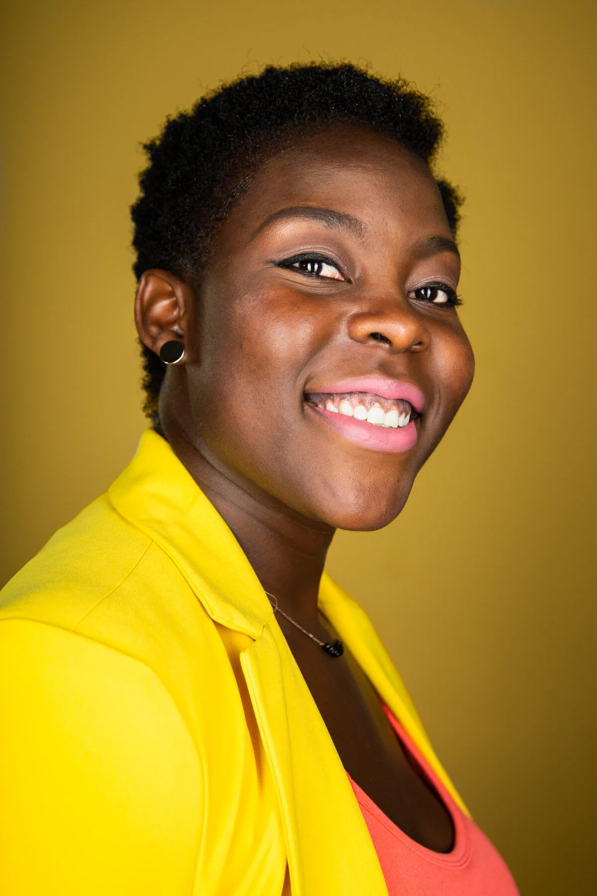 Smiling African American woman with short black hair wearing a yellow blazer and coral top against a yellow background.