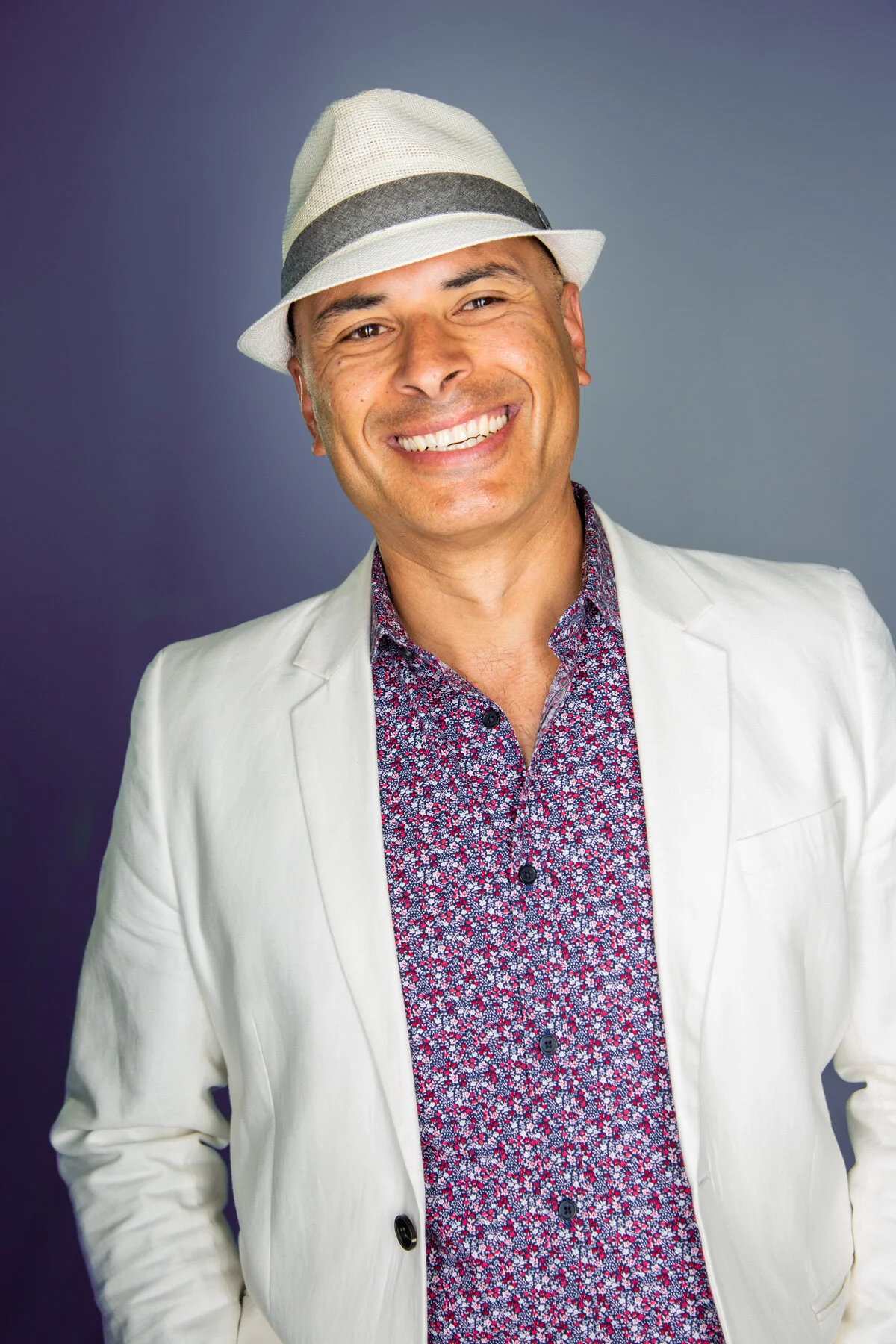 Smiling man in white blazer, patterned shirt, and white fedora hat against a gradient background.