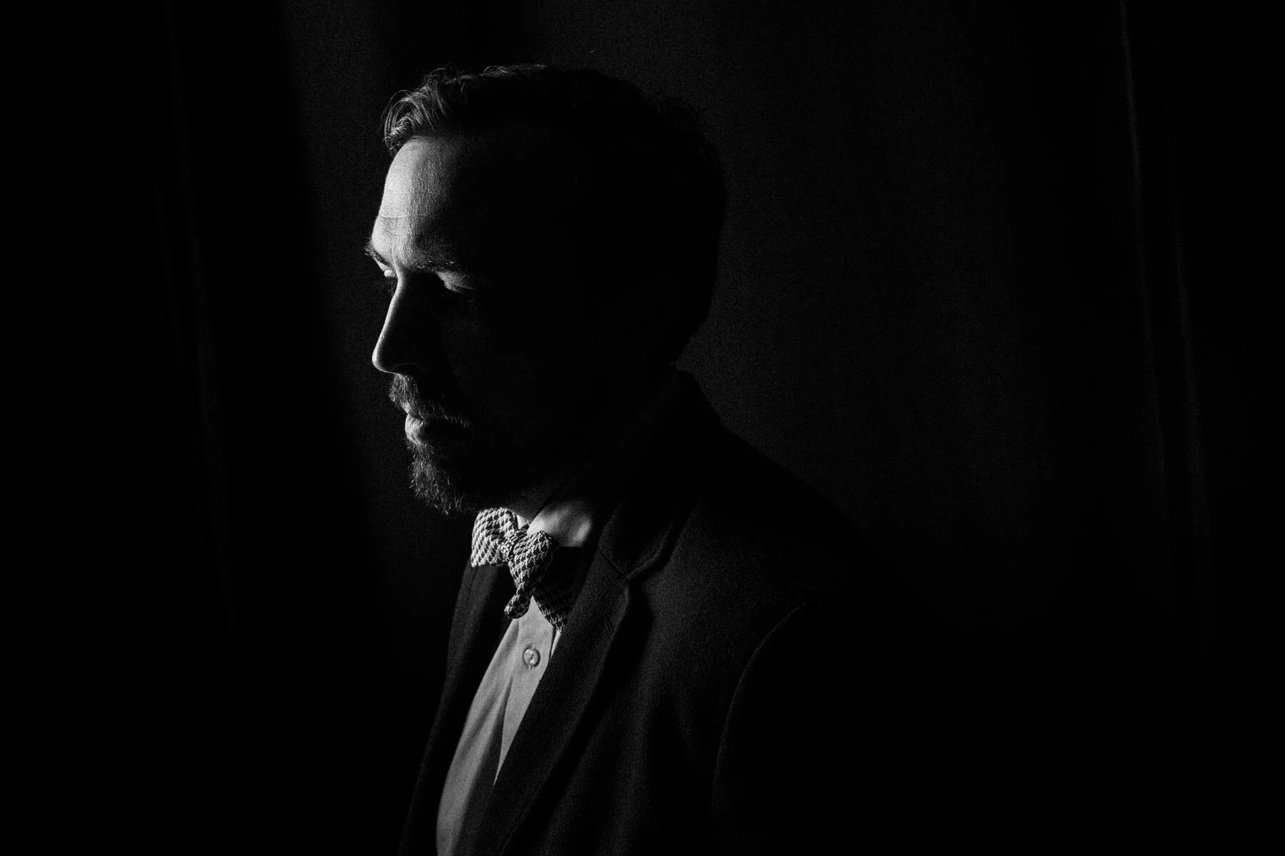 Black and white portrait of a man with a beard, closed eyes, wearing a suit and bow tie, in profile, with shadows highlighting his facial features, against a dark background.