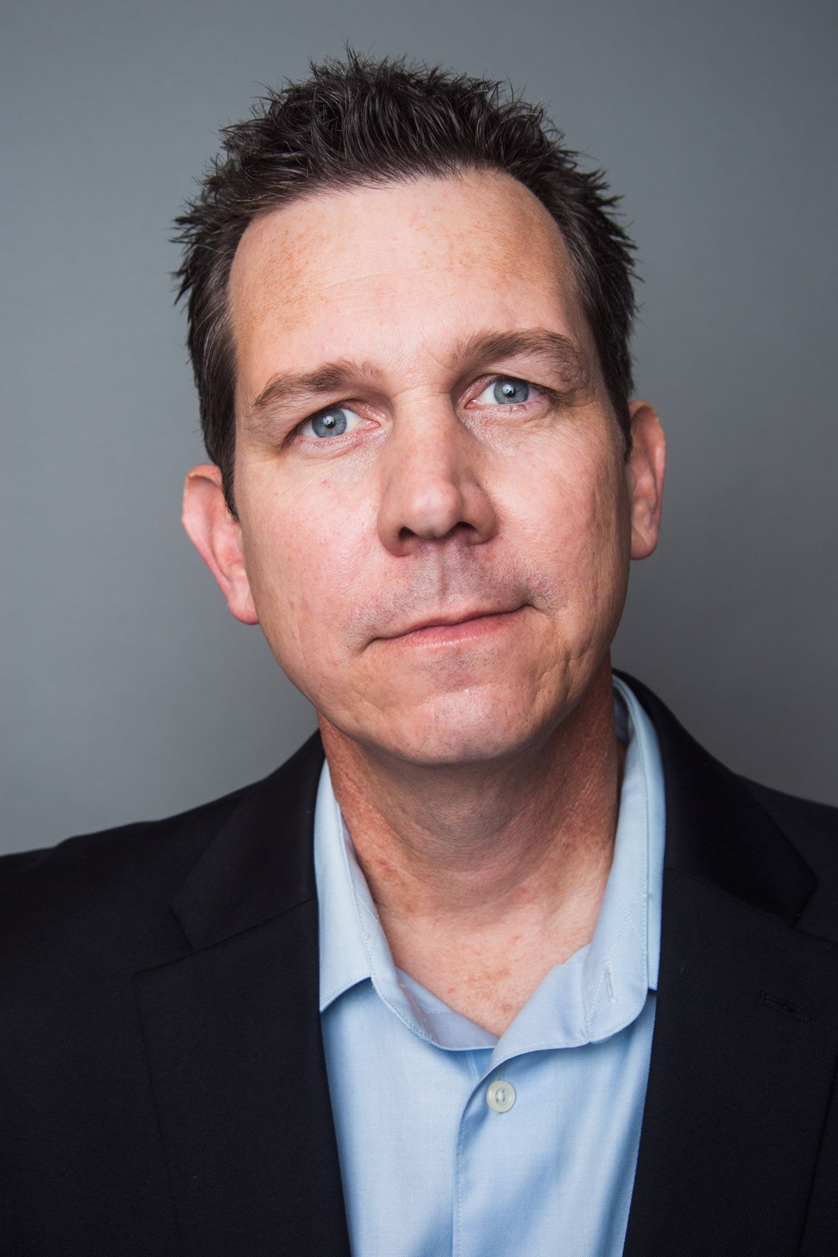 Portrait of a man with short dark hair, blue eyes, wearing a black suit jacket and a light blue collared shirt, against a plain grey background.