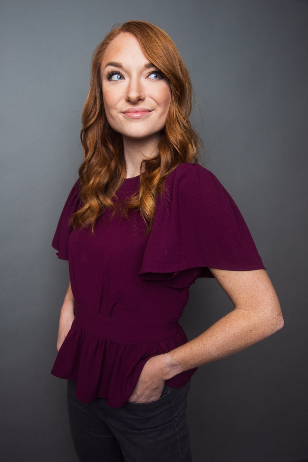 A woman with wavy red hair wearing a purple blouse standing against a gray background.