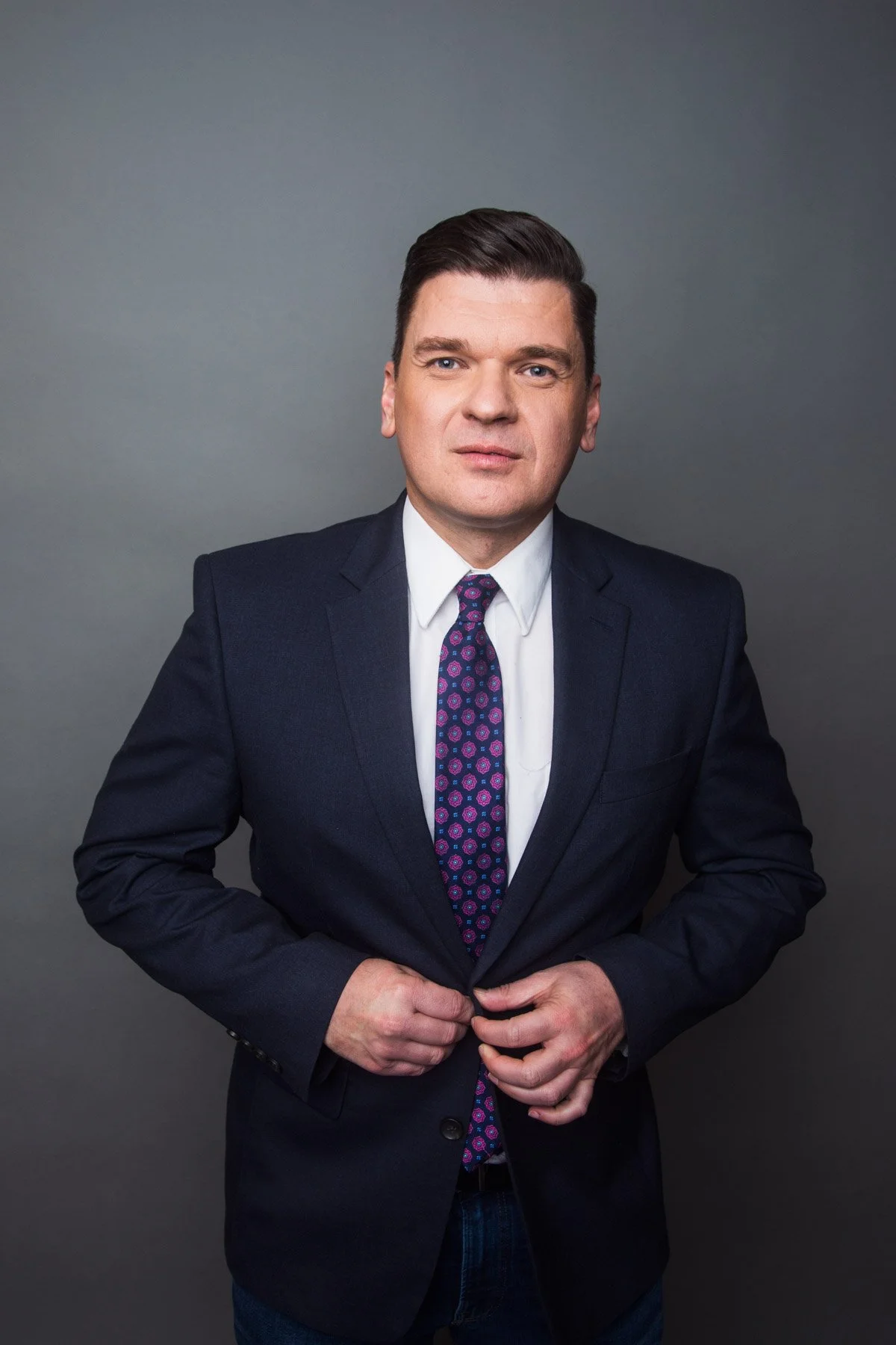 A man in a dark suit, white shirt, and patterned tie standing against a gray background. He is adjusting his suit jacket and looking at the camera.
