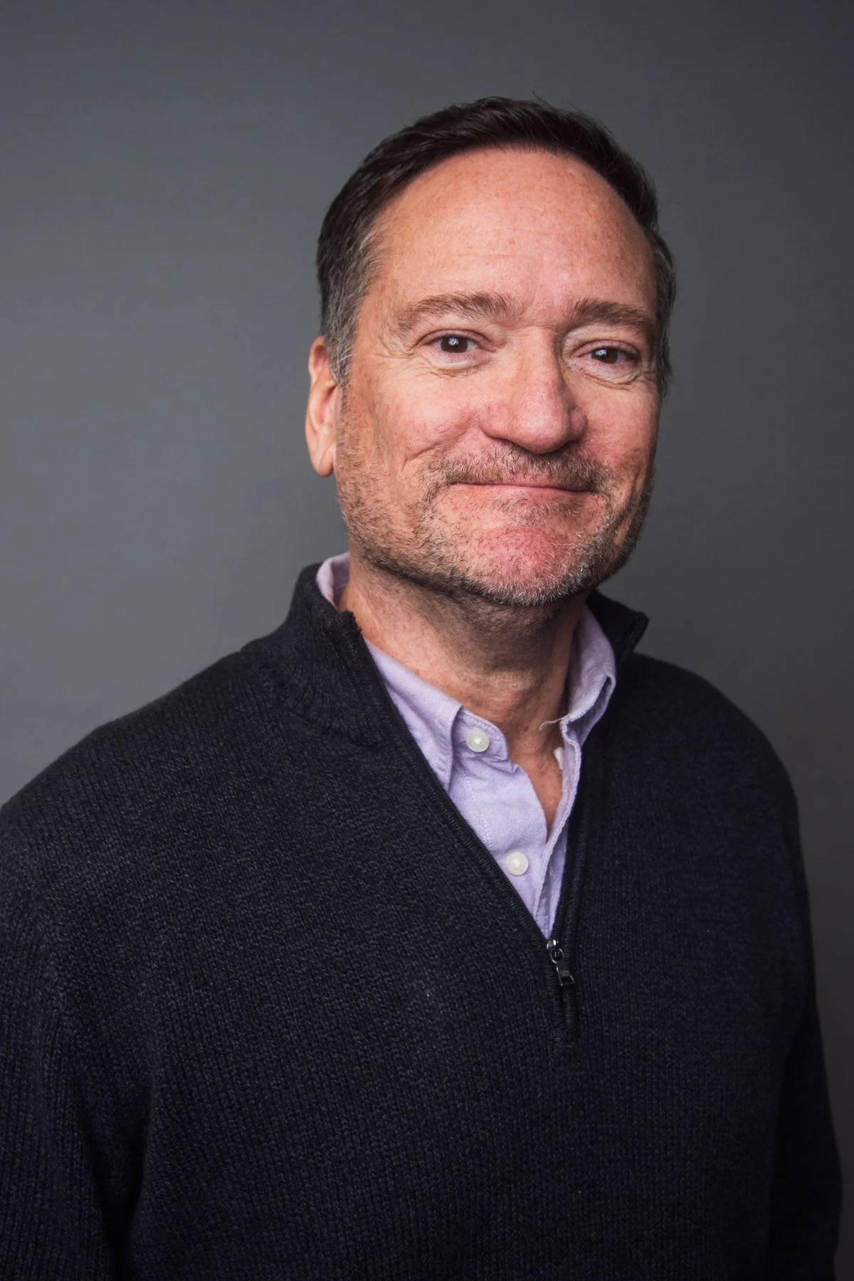 A middle-aged man with short dark hair, light skin, and a light beard, wearing a purple collared shirt underneath a black zip-up sweater, smiling slightly against a plain gray background.