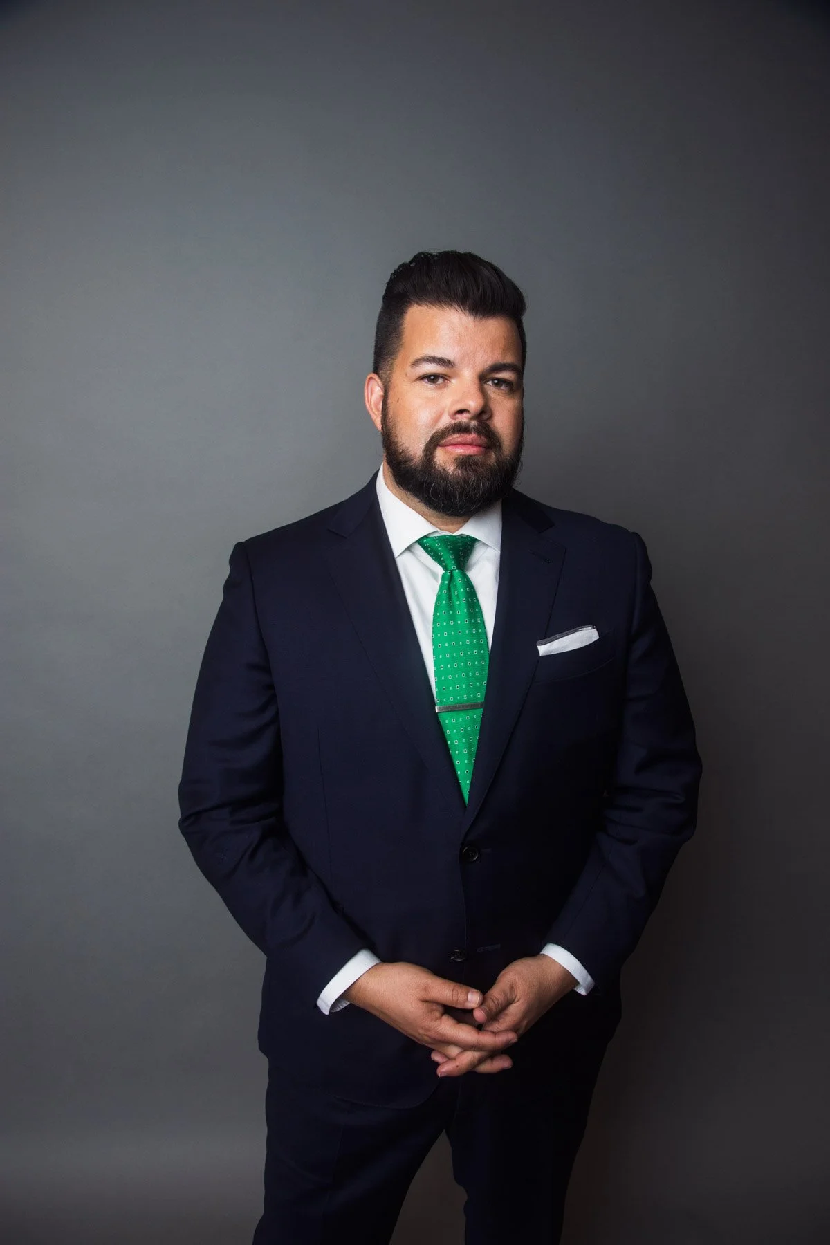 Man in dark suit with white shirt and green tie, standing against a plain gray background.