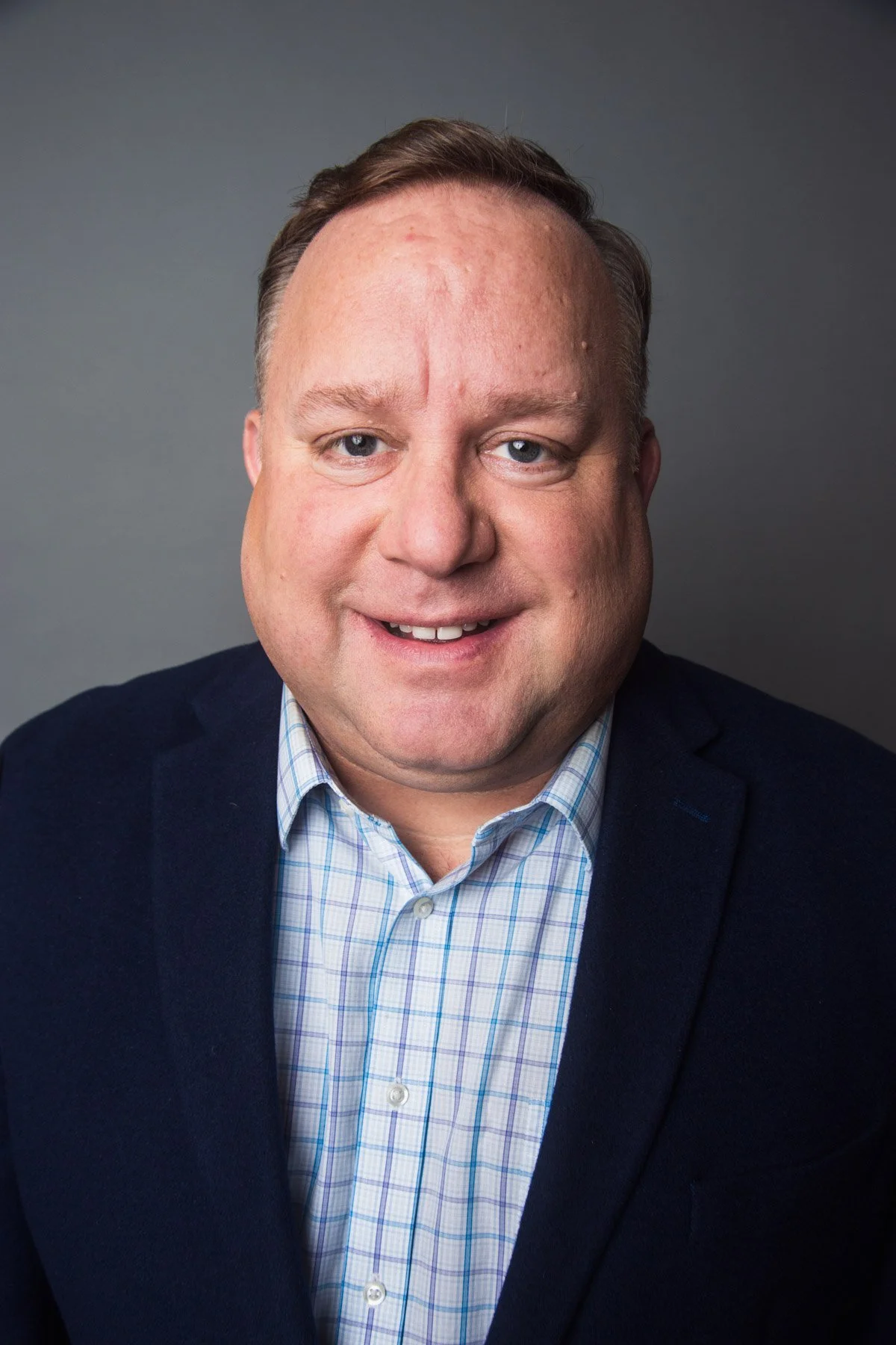 Professional headshot of a man wearing a dark blazer and a light blue checkered shirt, smiling in front of a gray background.