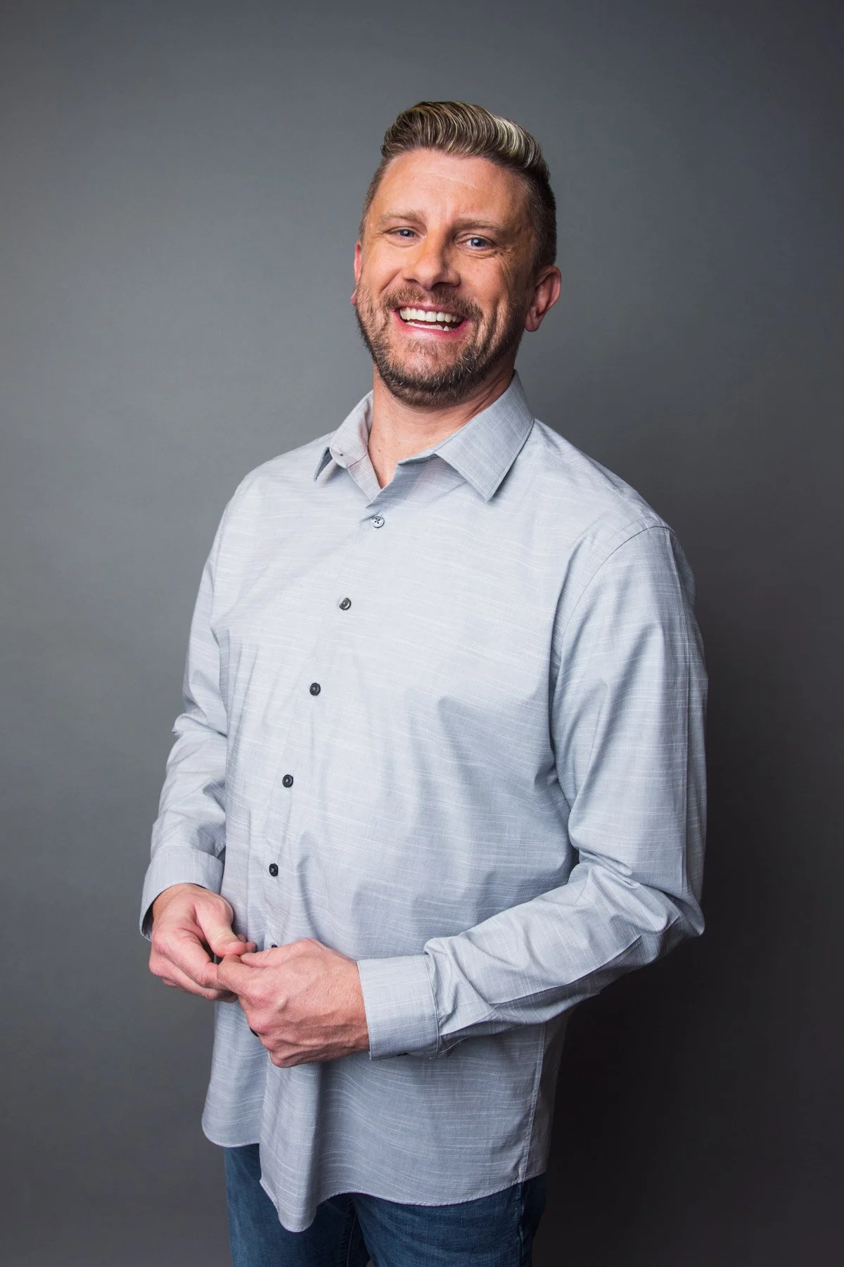 A smiling man with short light hair and a beard, wearing a light gray button-up shirt, standing against a dark gray background.