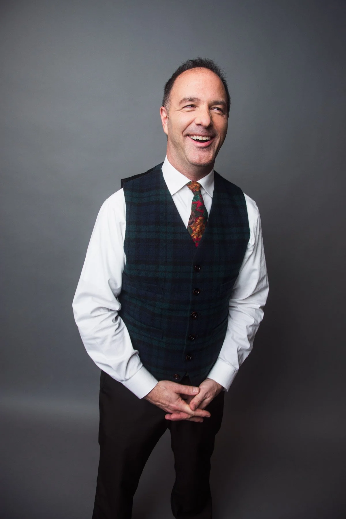 A man standing against a gray background, smiling and wearing a plaid vest over a white shirt with a patterned tie.