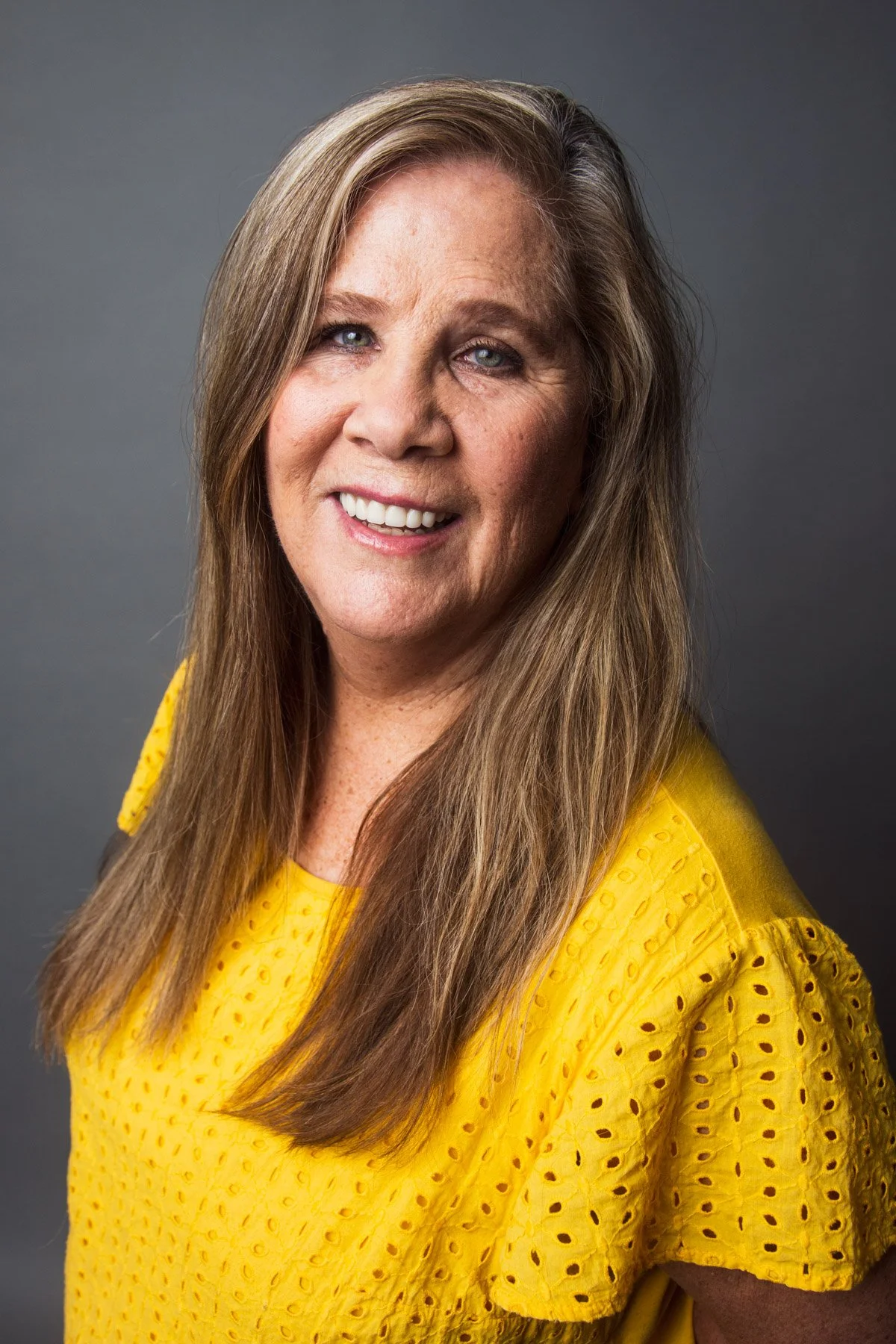 A woman with long light brown hair, blue eyes, and a friendly smile, wearing a yellow blouse with eyelet details, posing against a gray background.