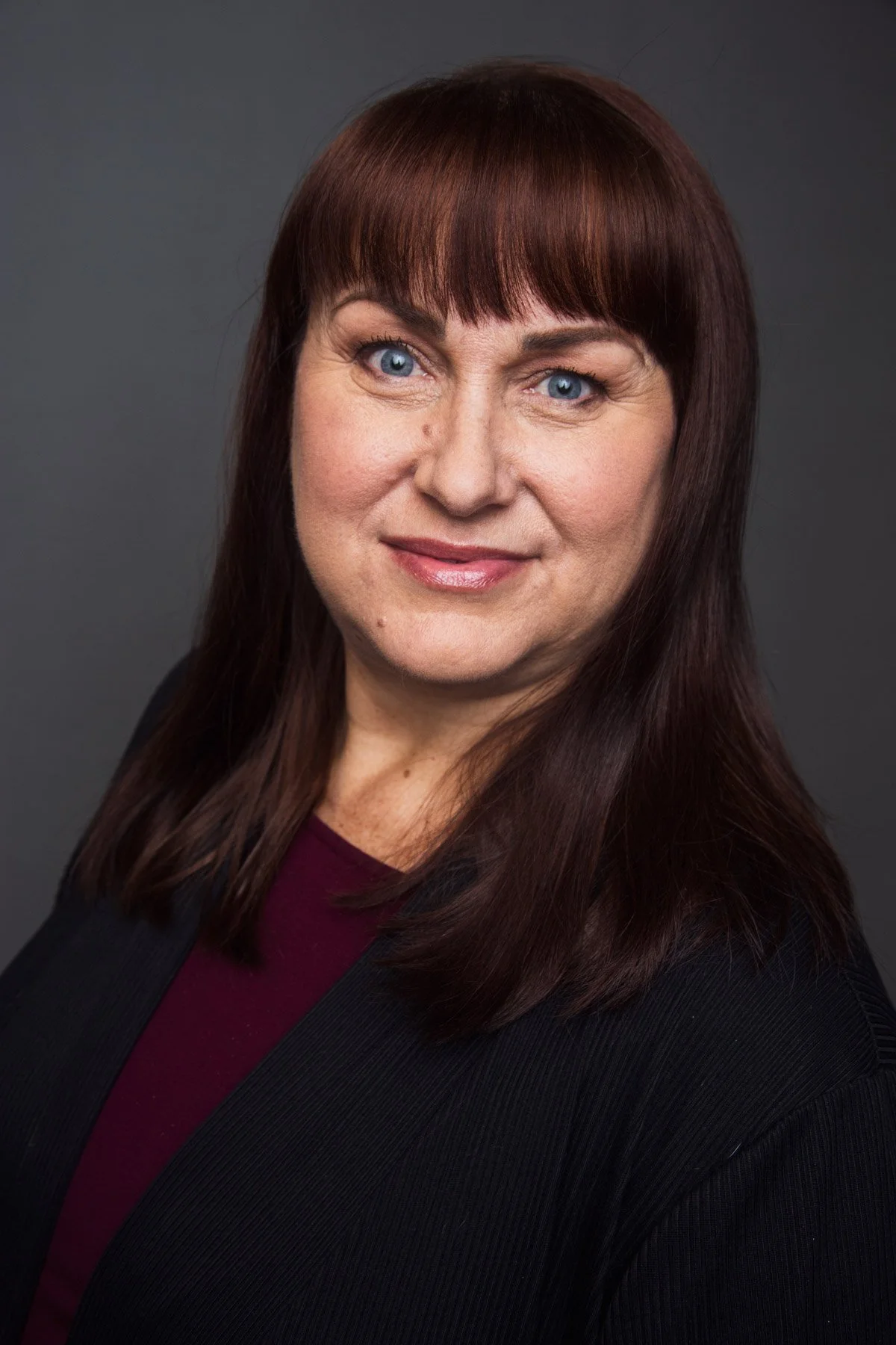 Headshot of a woman with shoulder-length dark red hair, blue eyes, and light skin, wearing a black blazer over a maroon top, posed against a dark gray background.