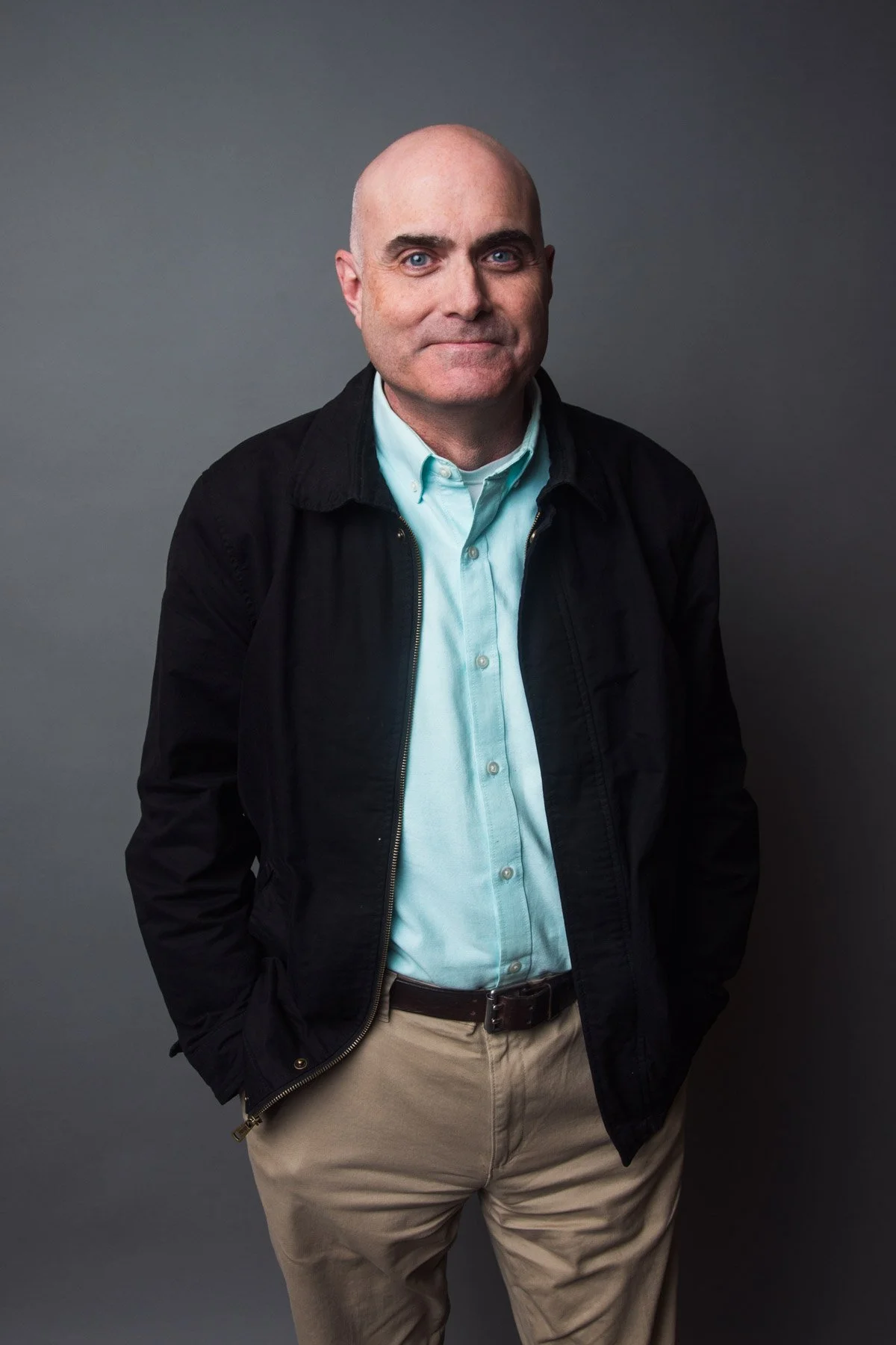 A bald man wearing a light blue button-up shirt, black jacket, and khaki pants posing against a gray background.