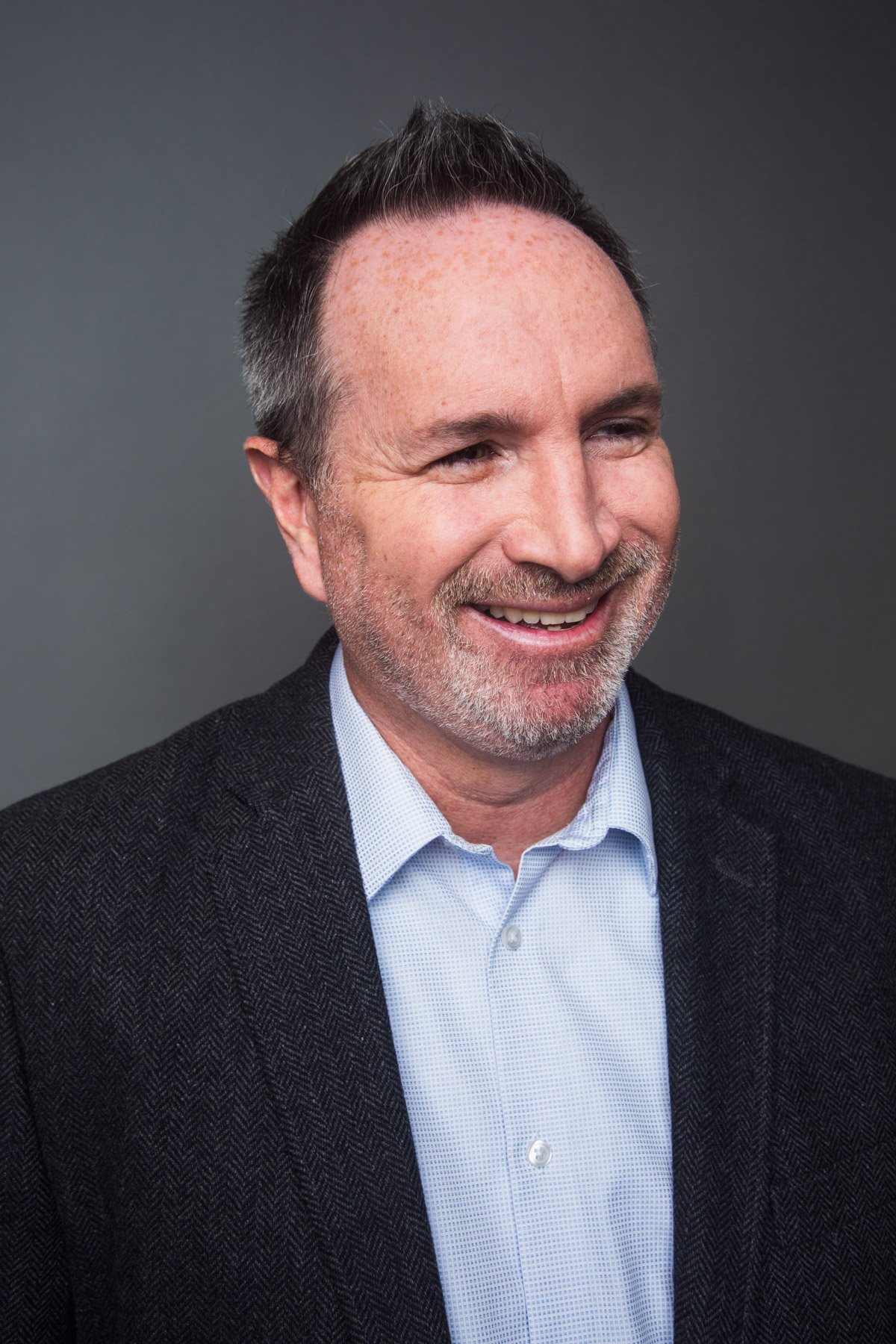 A middle-aged man with short, styled dark hair with gray, smiling, wearing a light blue shirt and a dark textured blazer, standing against a plain dark background.