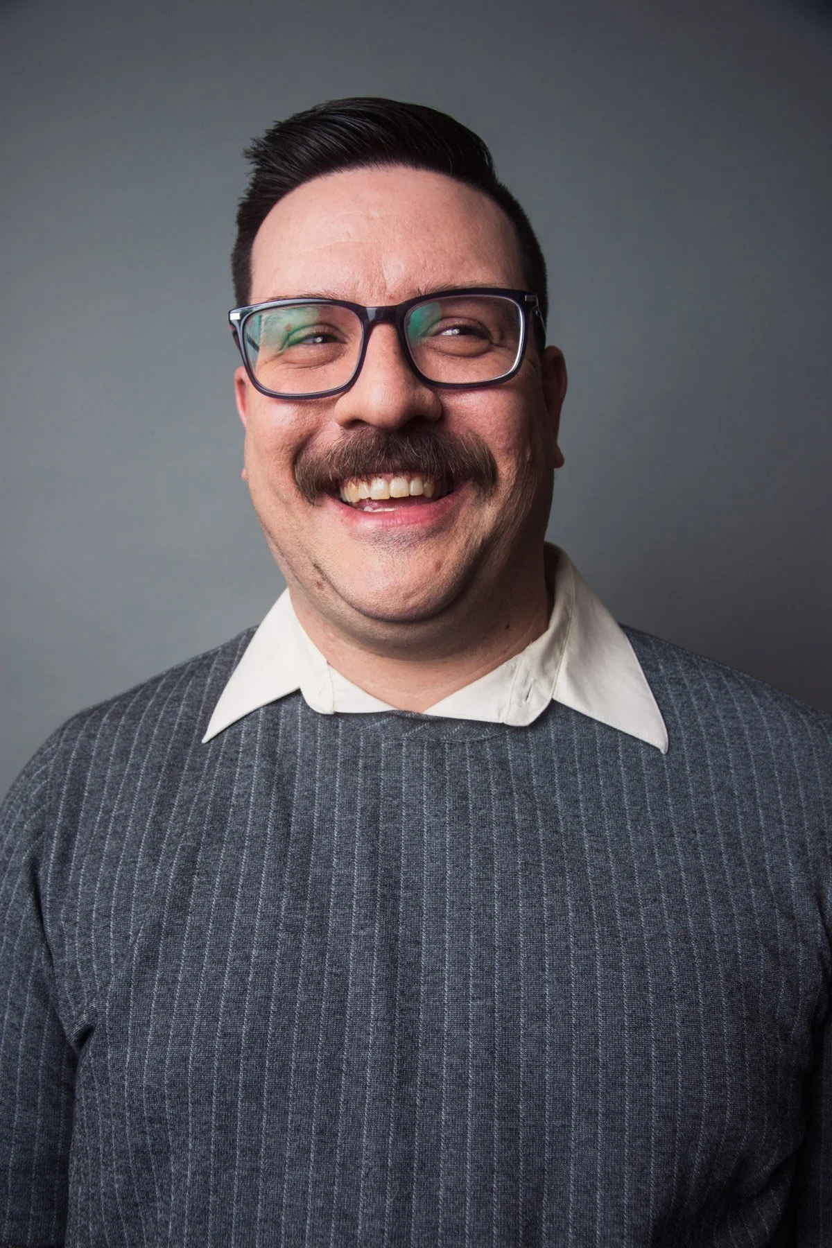 A man with short dark hair, glasses, a moustache, and a big smile, wearing a white collared shirt and a dark grey sweater with thin vertical stripes, standing against a plain grey background.