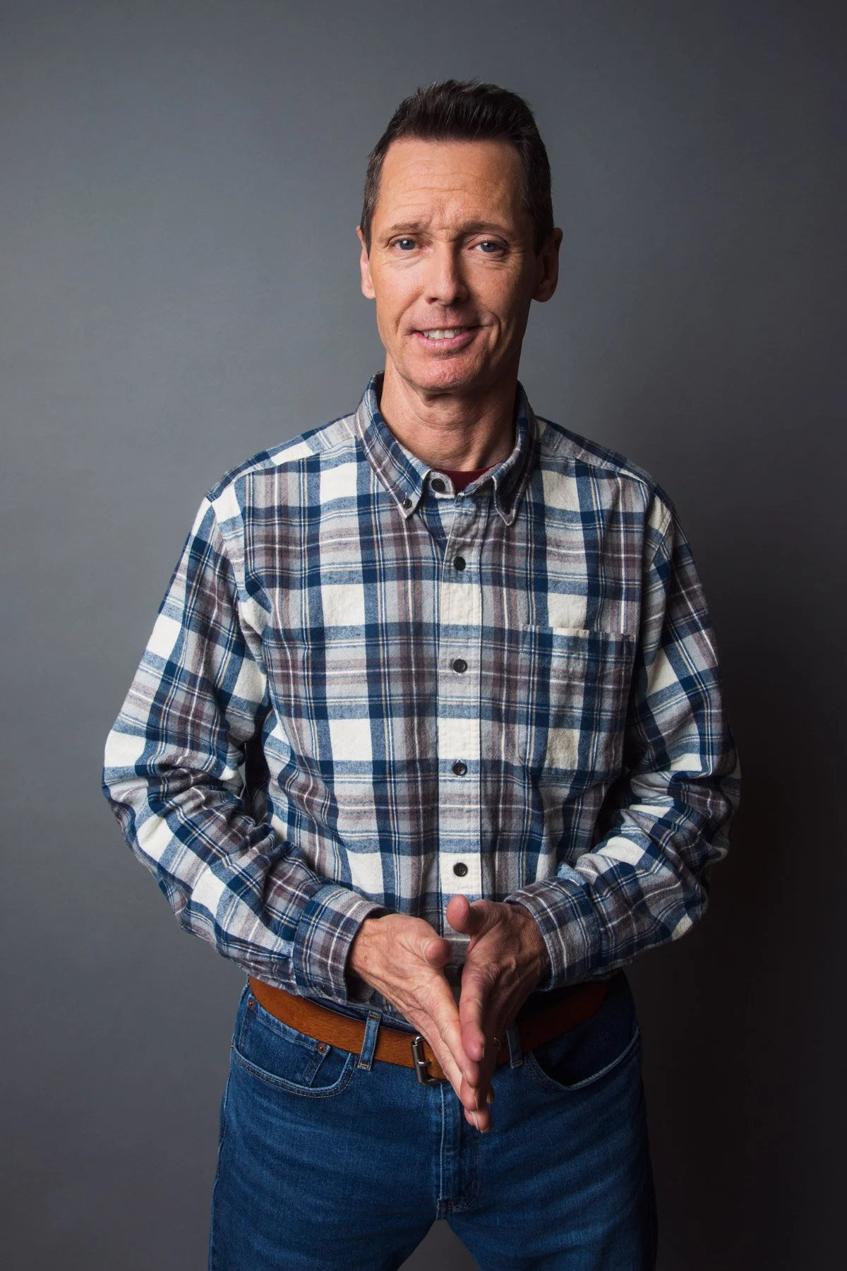 A middle-aged man with short dark hair, wearing a plaid shirt and jeans, standing against a gray backdrop, with one eyebrow raised and a slight smile.