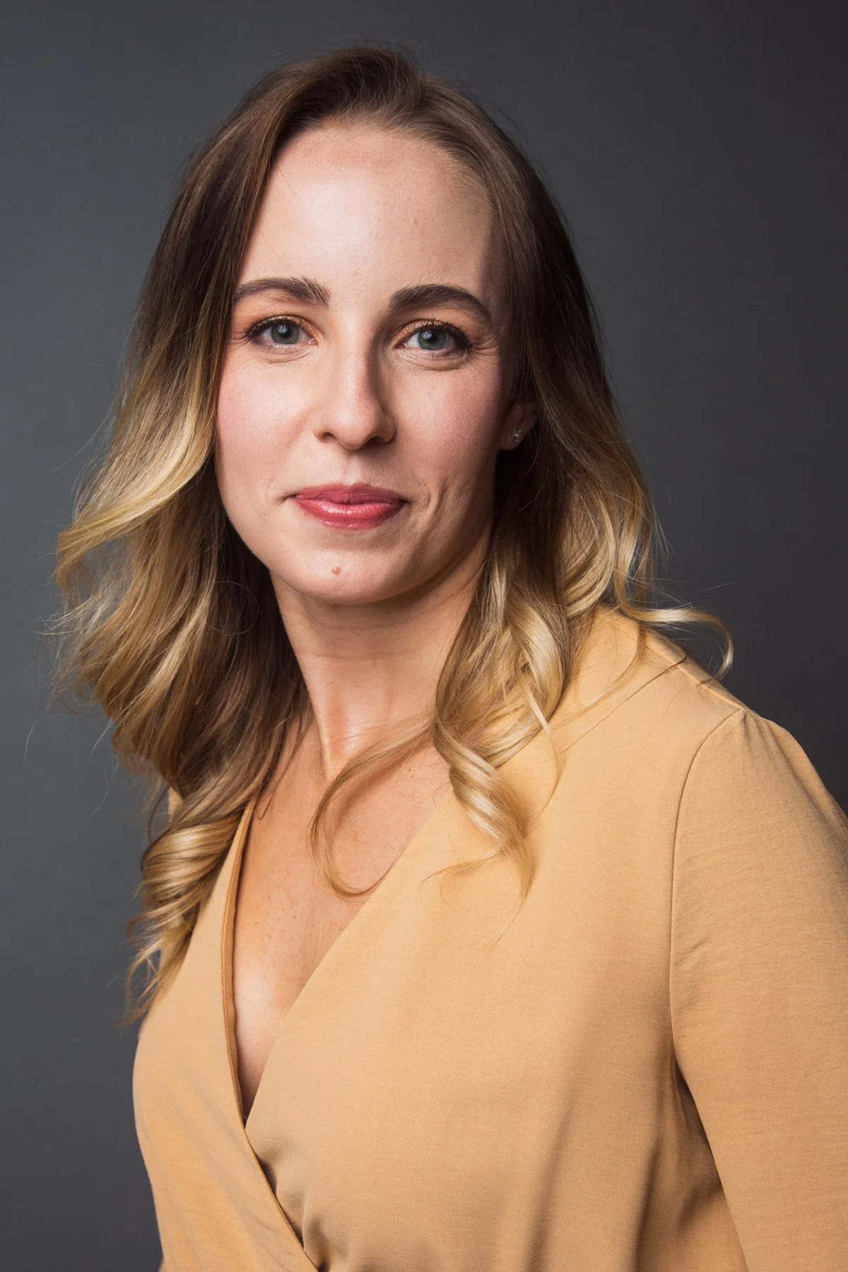 A portrait of a woman with blonde hair styled in loose waves, wearing a tan blouse, against a dark gray background.
