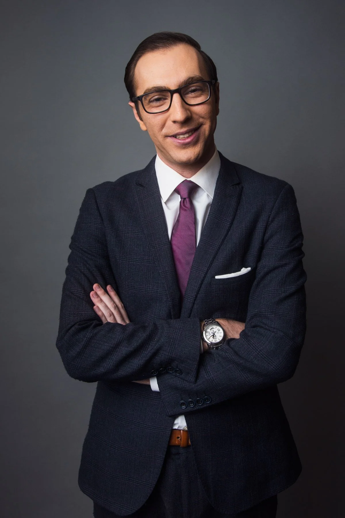 A man in a dark suit with a purple tie, wearing glasses, smiling with arms crossed, against a gray background.