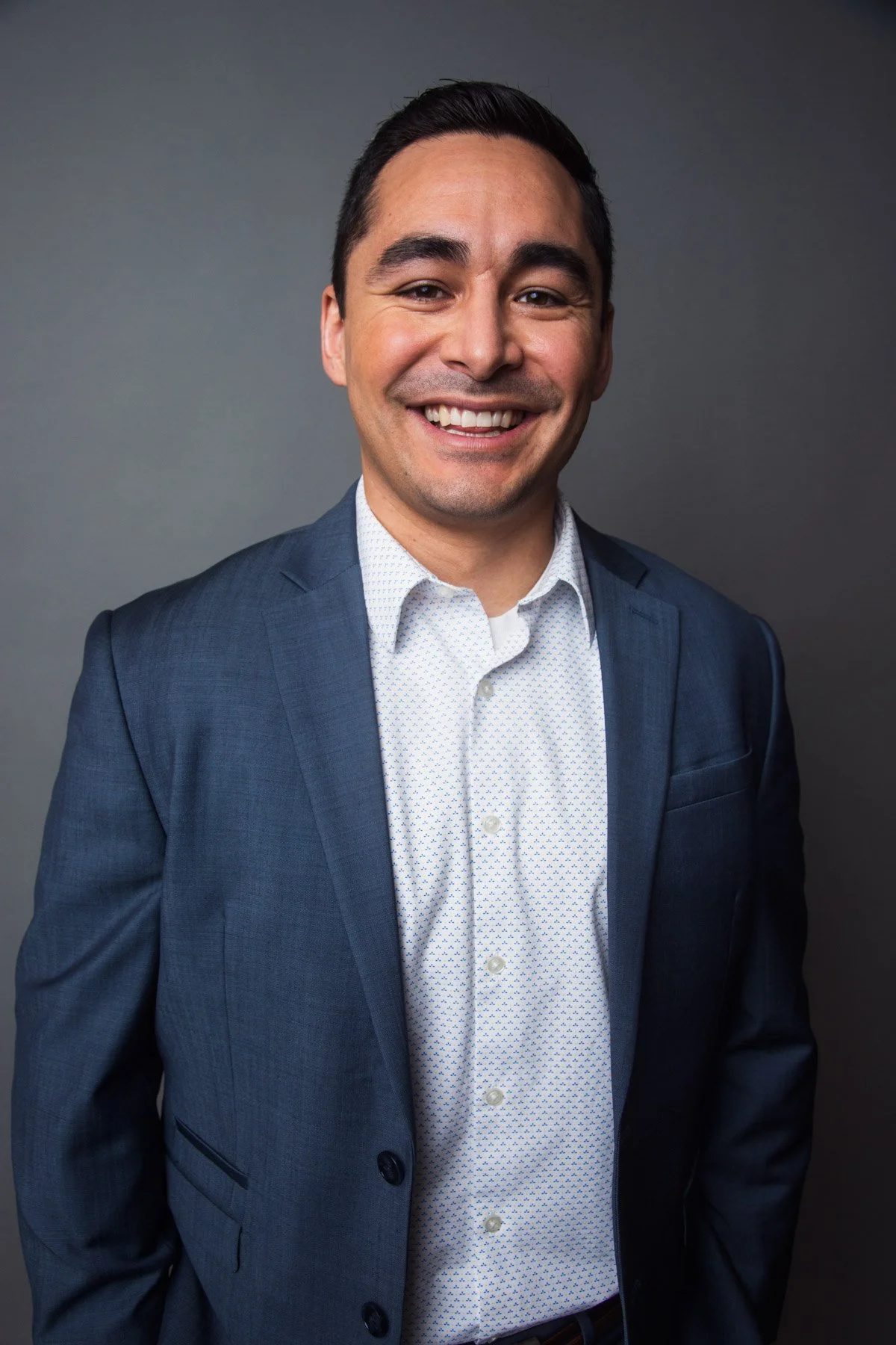 Portrait of a smiling man in a navy blue suit and white shirt standing against a gray background.