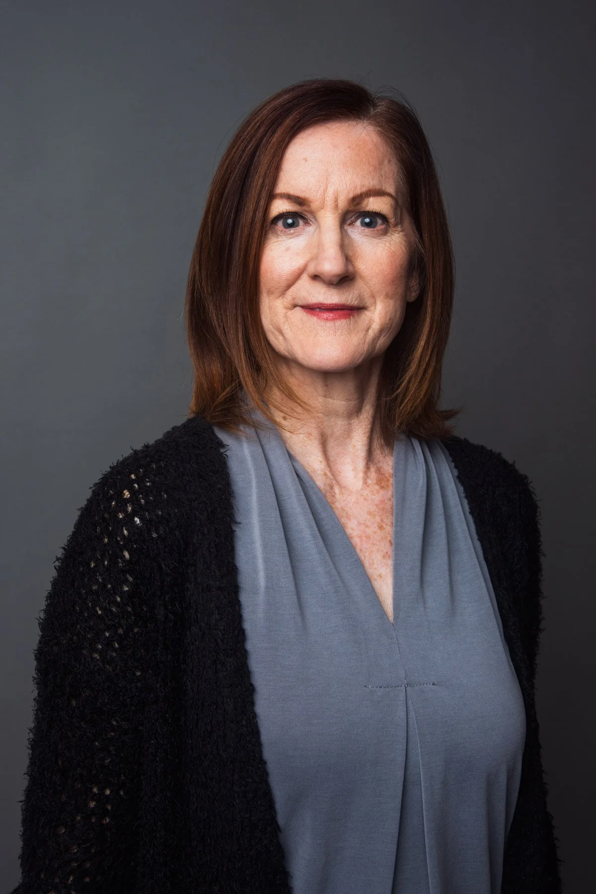 A woman with shoulder-length reddish-brown hair, wearing a gray top and black knitted cardigan, standing against a gray background.