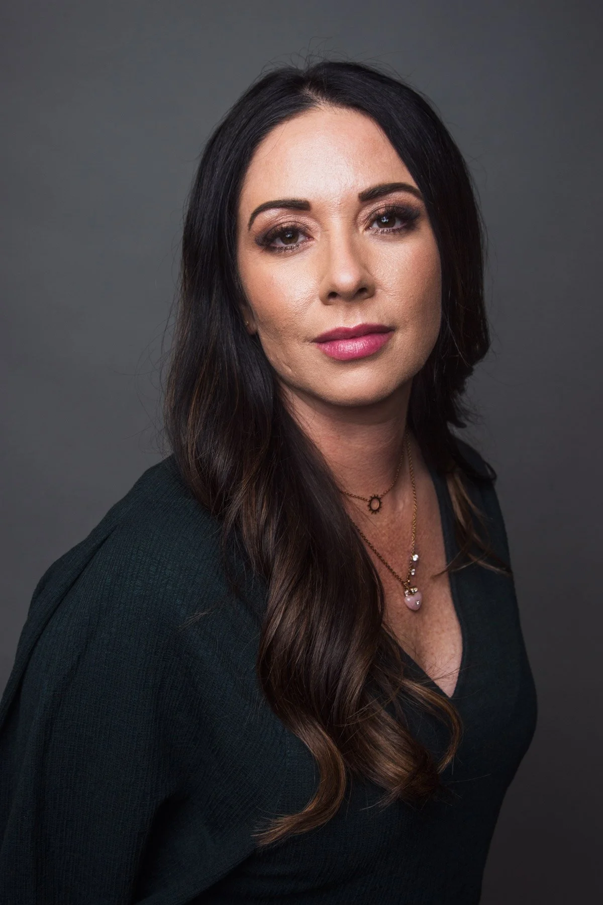 Portrait of a woman with long dark hair, wearing a black top and layered necklaces, against a plain gray background.