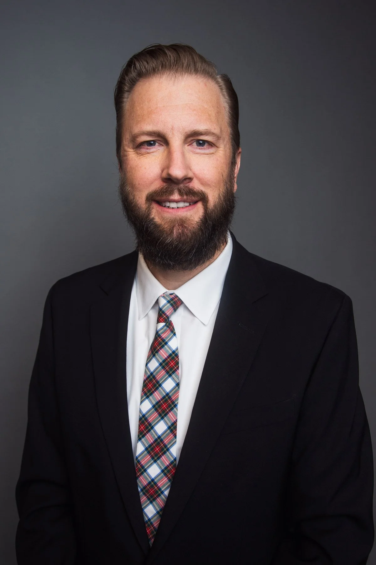 A man with a beard and mustache in a black suit, white shirt, and colorful plaid tie, smiling against a gray background.